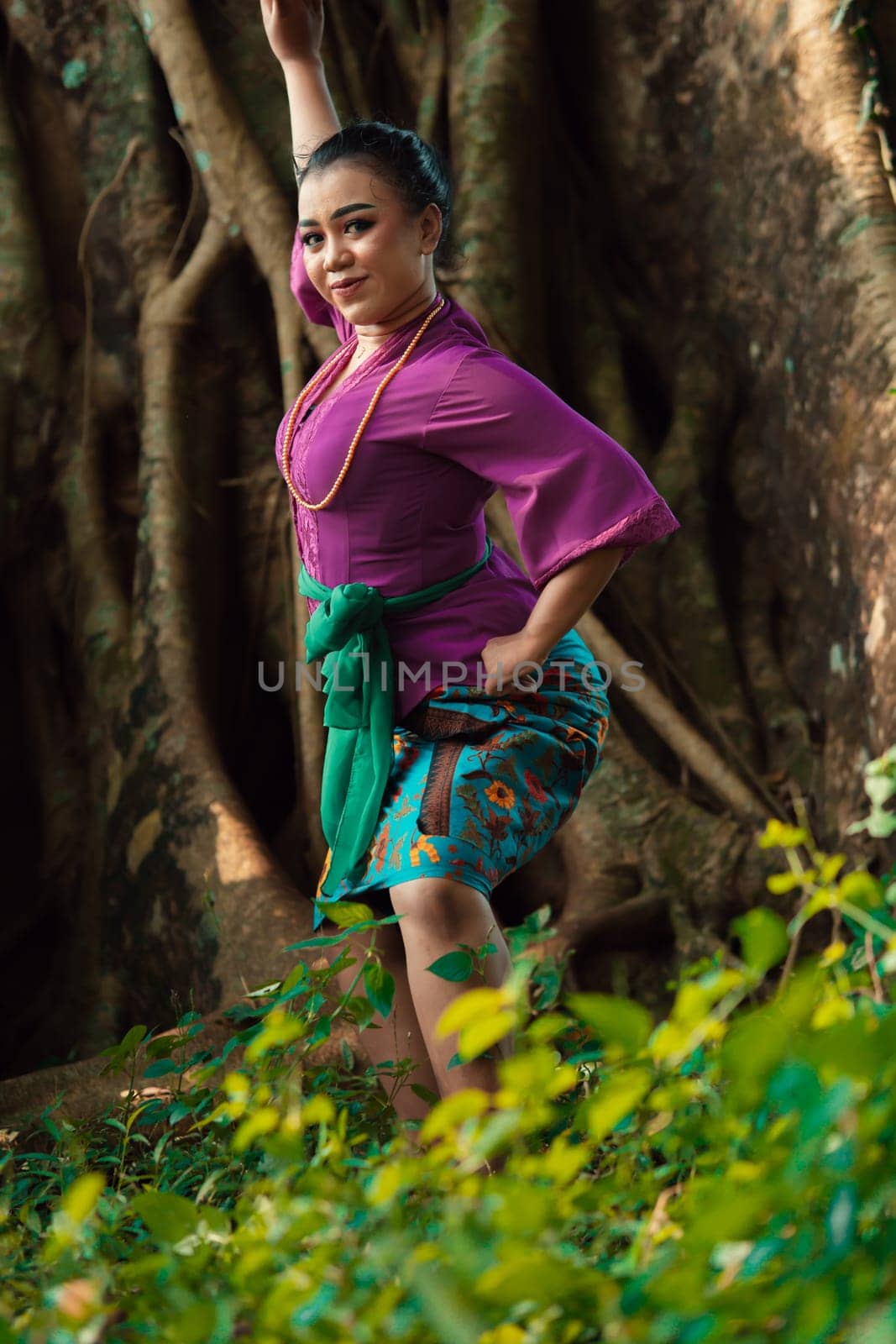 Asian woman takes a dancing pose in the jungle while wearing a purple dress and a golden necklace with a beautiful smile on her face
