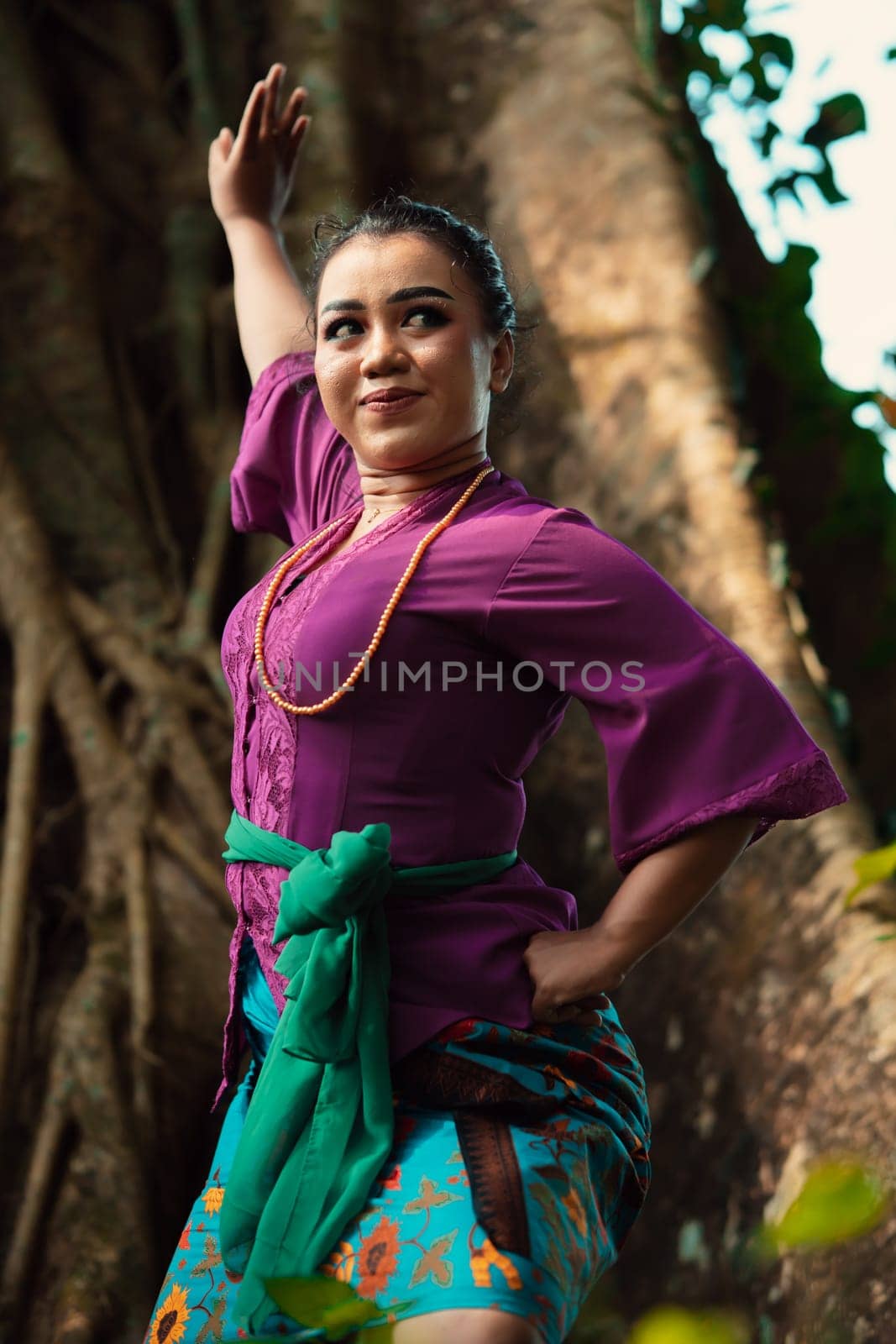 An Indonesian woman bravely poses while raising her hands in a purple dress while standing in the forest