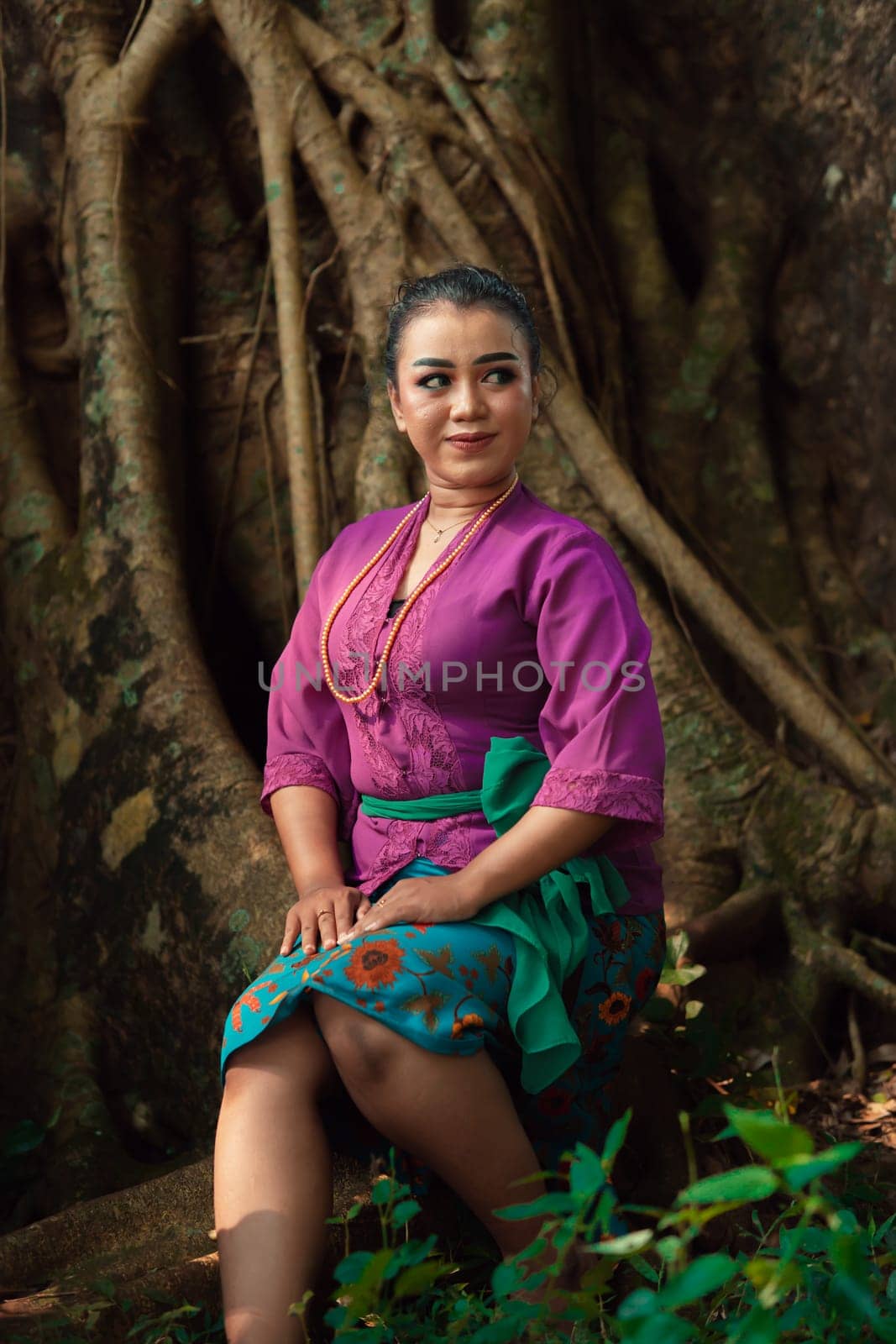An Asian woman beautifully sitting on the big tree in a purple dress while visiting the jungle in the daylight