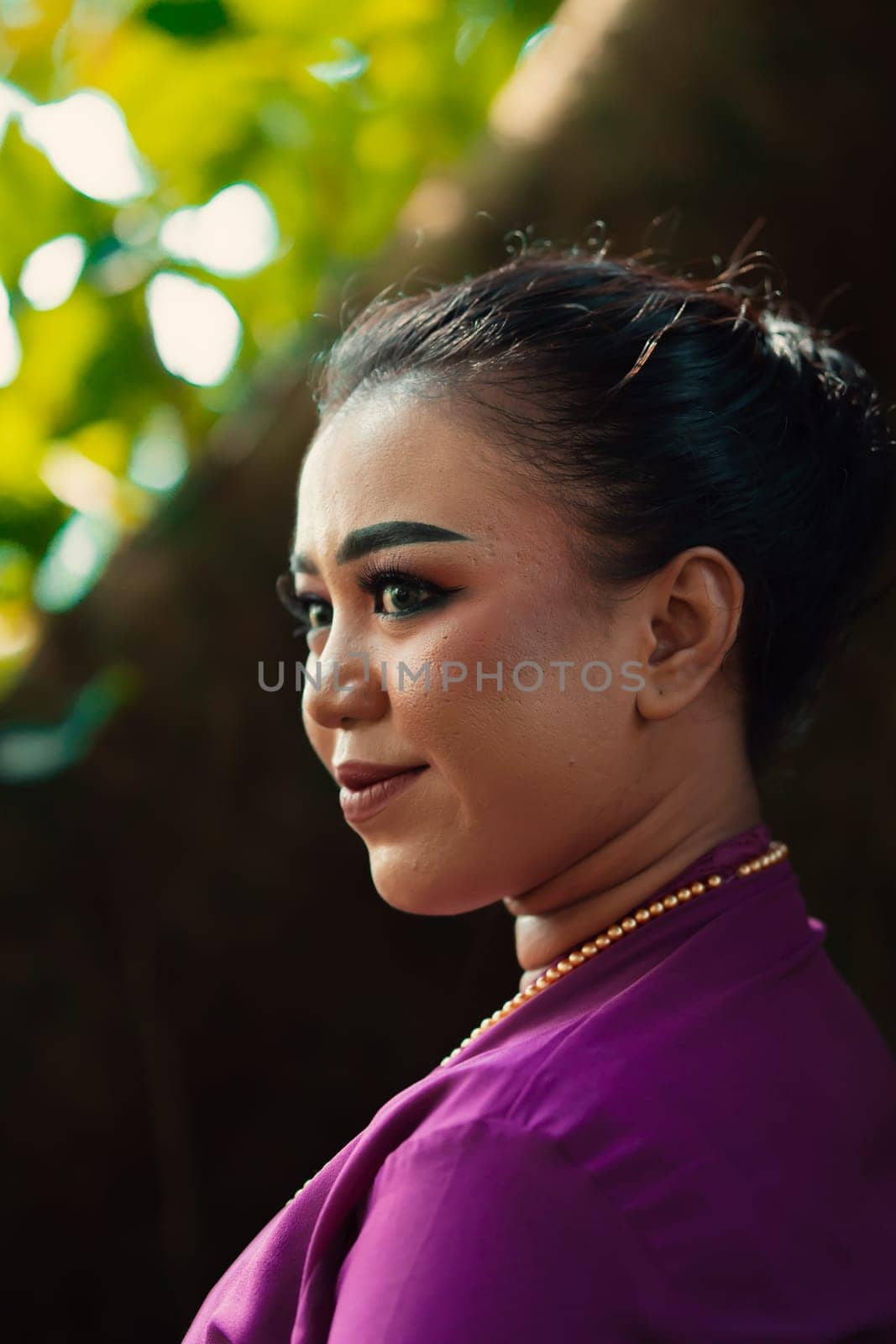 Indonesian woman smiling in a traditional purple dress while wearing makeup and golden jewelry in front of the big tree inside the forest