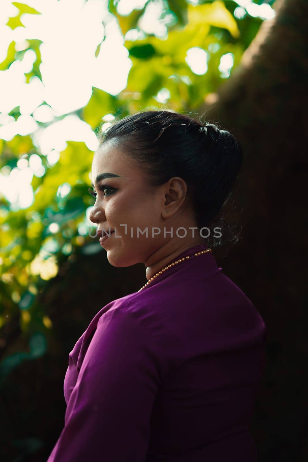 Indonesian woman smiling in a traditional purple dress while wearing makeup and golden jewelry in front of the big tree inside the forest