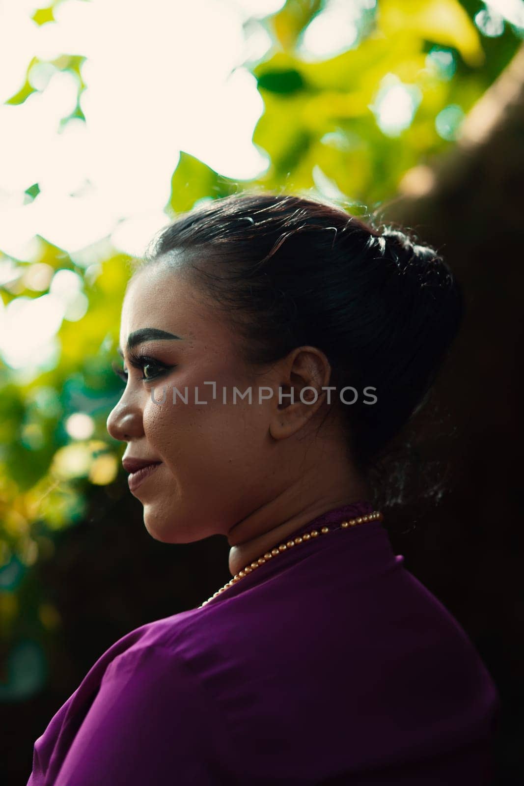 Indonesian woman smiling in a traditional purple dress while wearing makeup and golden jewelry in front of the big tree inside the forest