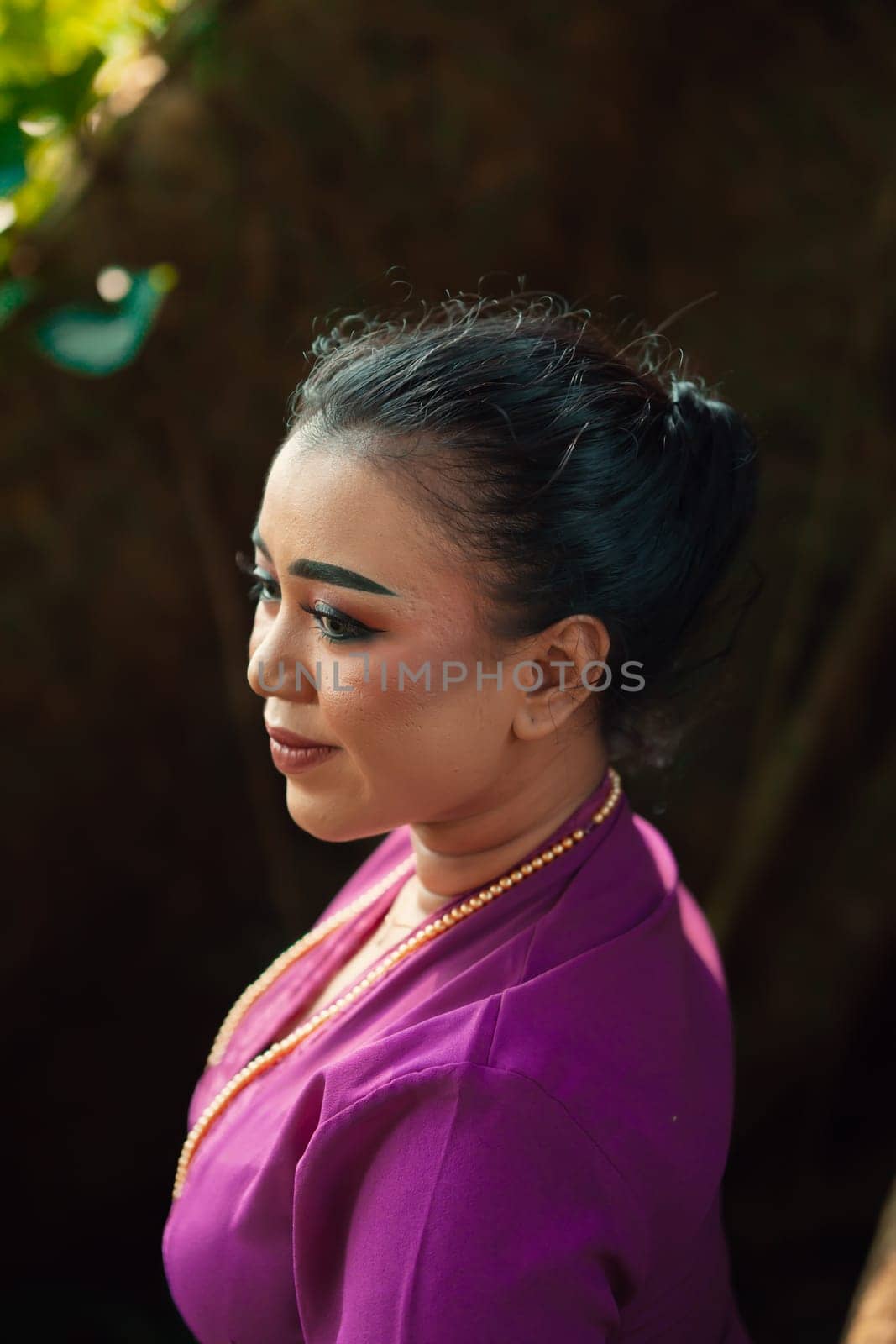 Indonesian woman smiling in a traditional purple dress while wearing makeup and golden jewelry in front of the big tree inside the forest