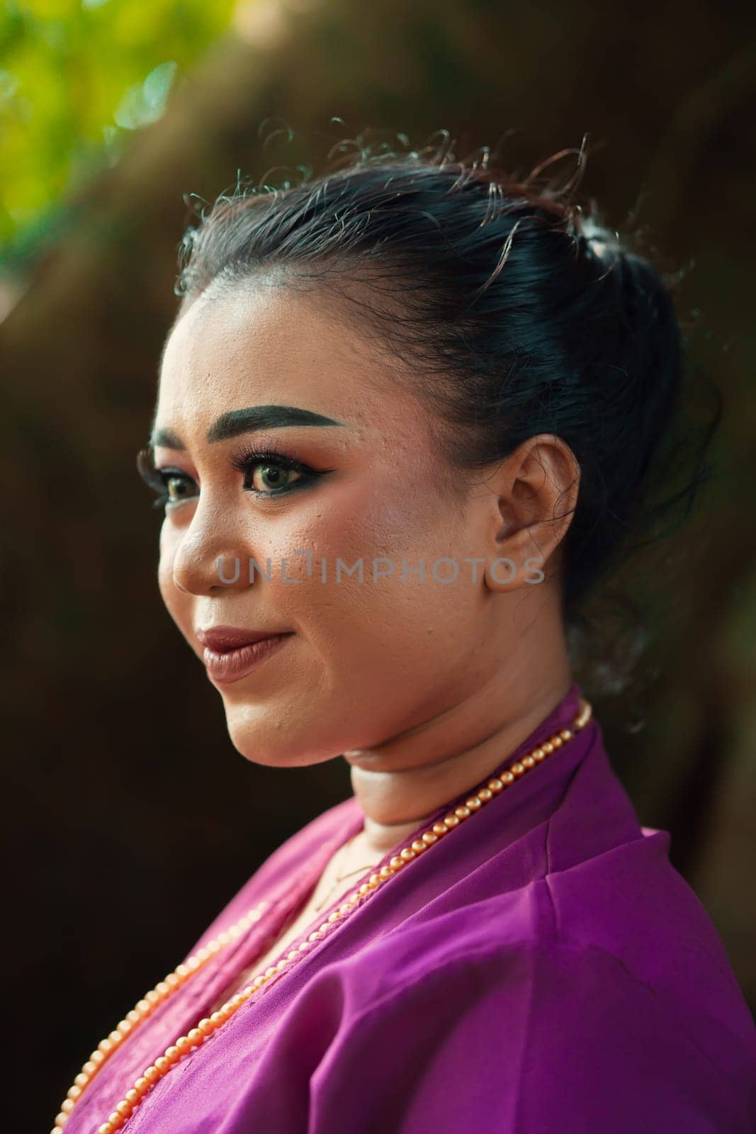 Indonesian woman smiling in a traditional purple dress while wearing makeup and golden jewelry in front of the big tree inside the forest