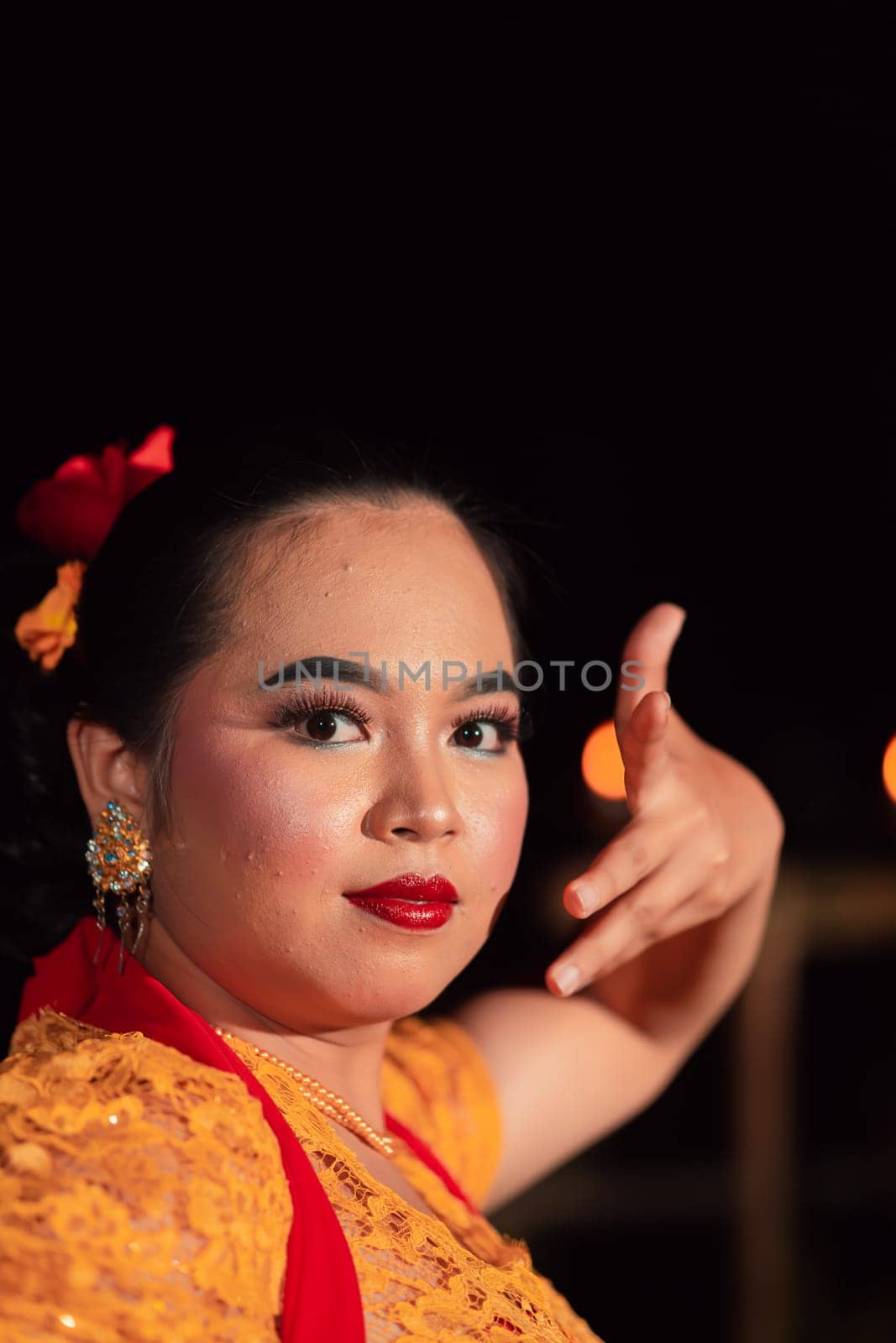 Balinese woman with red lips and makeup wearing a yellow dress and red scarf on a dark night inside the village