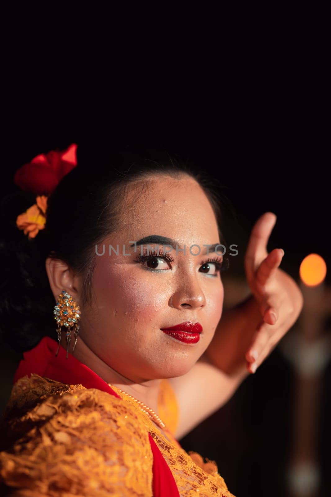 Balinese woman with red lips and makeup wearing a yellow dress and red scarf on a dark night inside the village