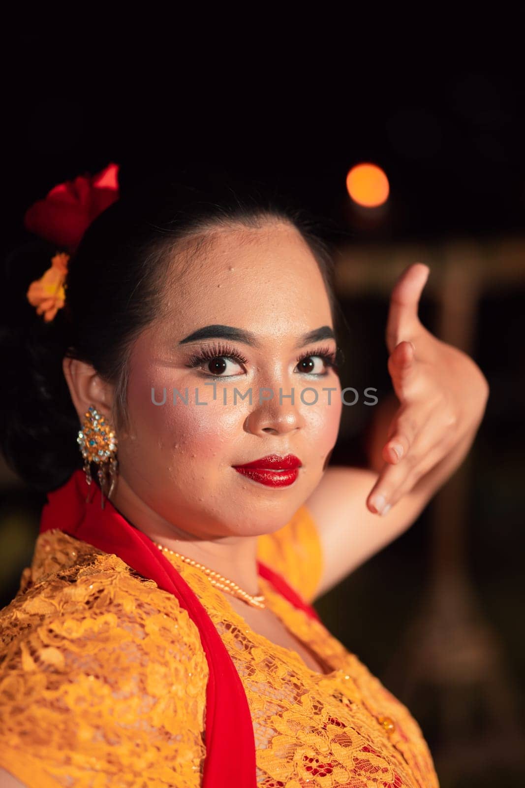 Balinese woman with red lips and makeup wearing a yellow dress and red scarf on a dark night inside the village