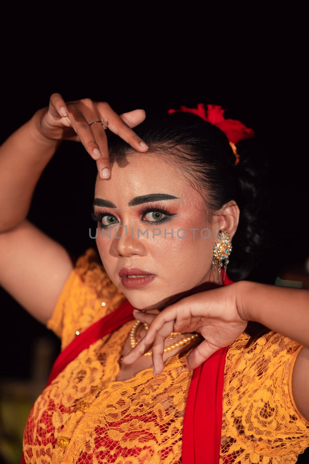 the fierce face of a Balinese woman with makeup in a traditional orange dress and red scarf while dancing at the festival in the night
