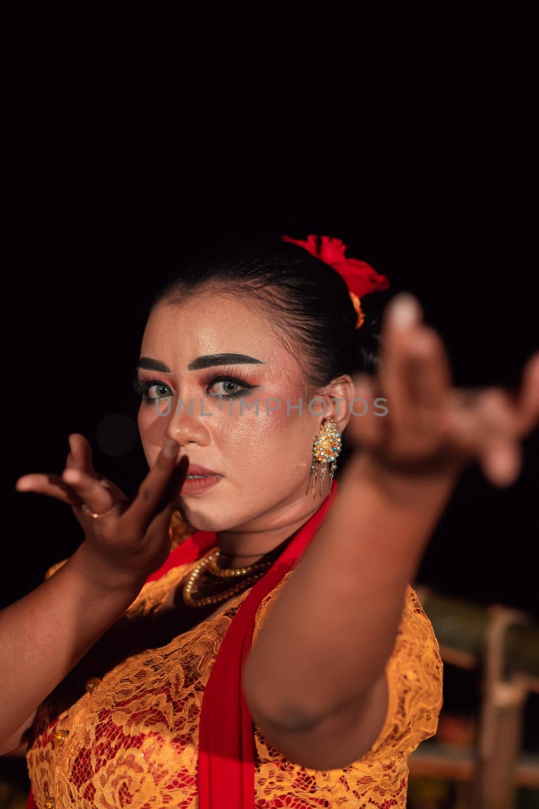 the fierce face of a Balinese woman with makeup in a traditional orange dress and red scarf while dancing at the festival in the night