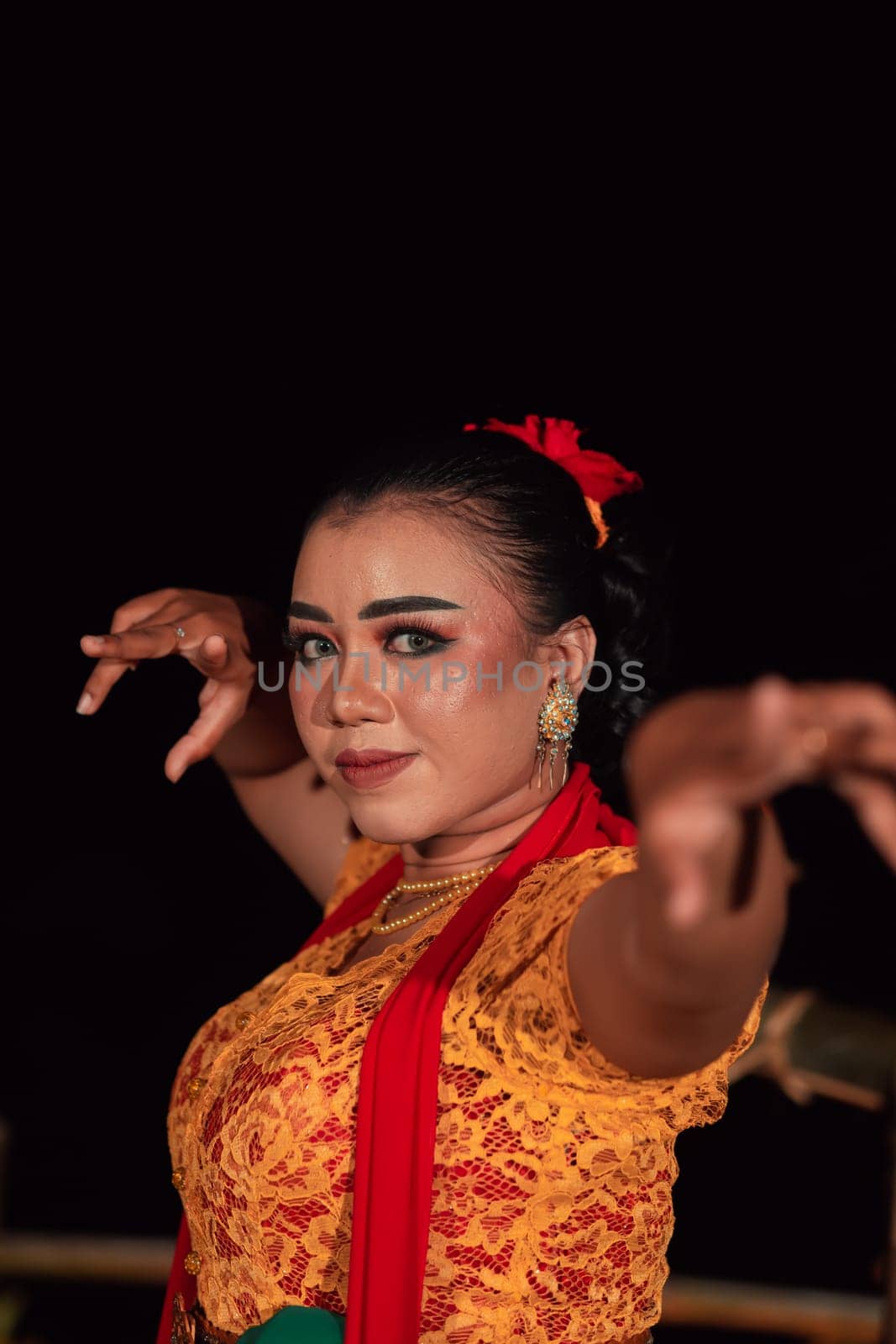 the fierce face of a Balinese woman with makeup in a traditional orange dress and red scarf while dancing at the festival in the night