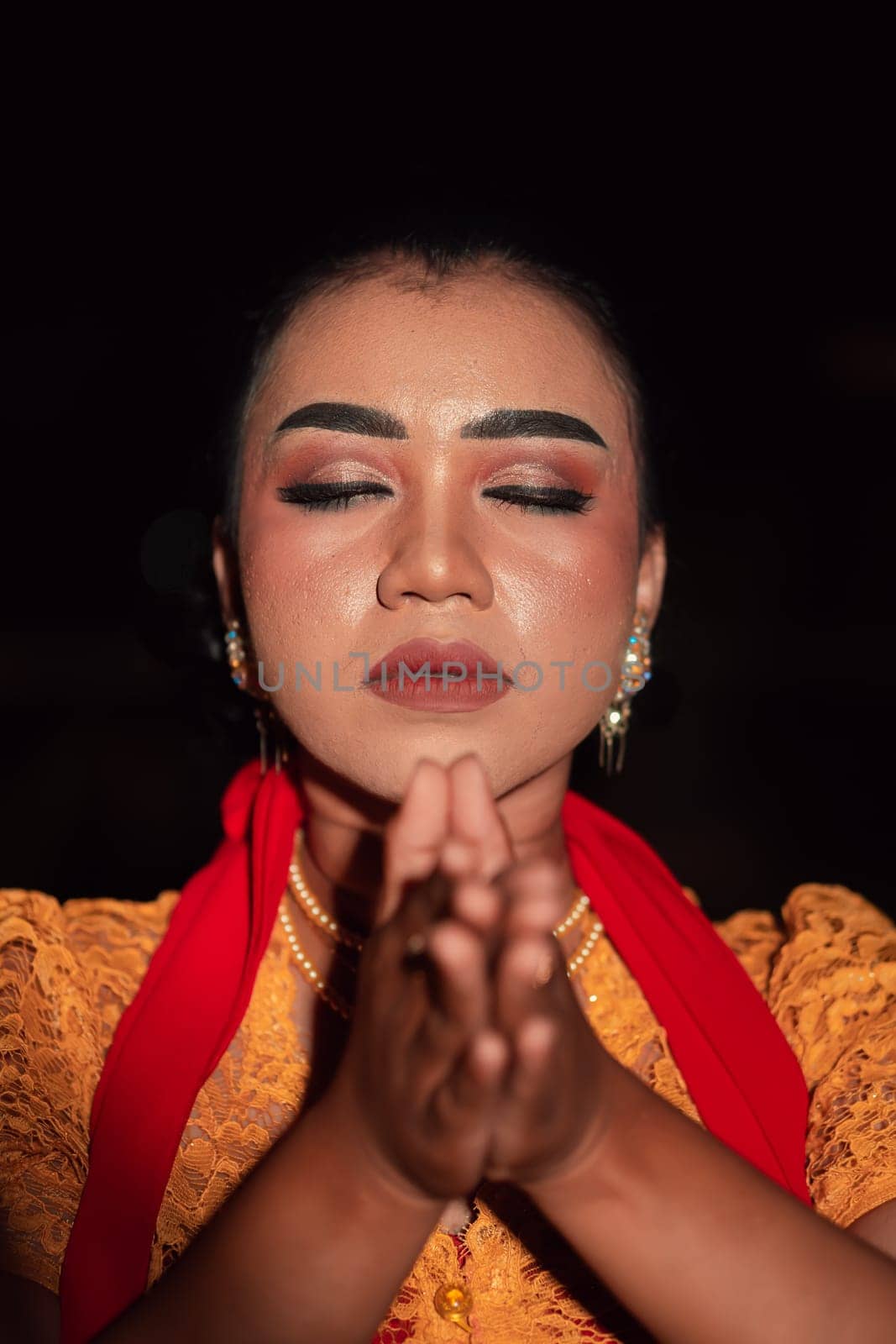 Asian woman praying with both her hands while wearing makeup and an orange dress with a red scarf on her body in the dark night
