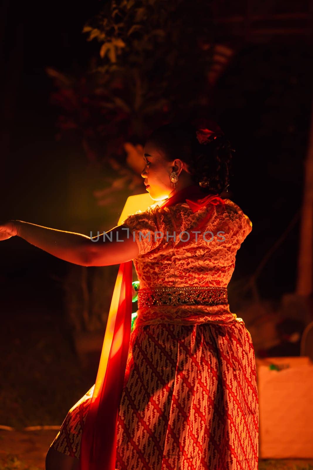 A Silhouette body of a Balinese woman in a traditional orange dress while dancing in front of the lighting on the dark night at the festival