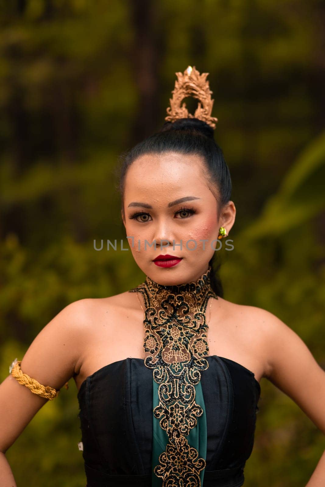 Balinese woman in makeup and gold jewelry on her body with a black costume standing in front of the jungle