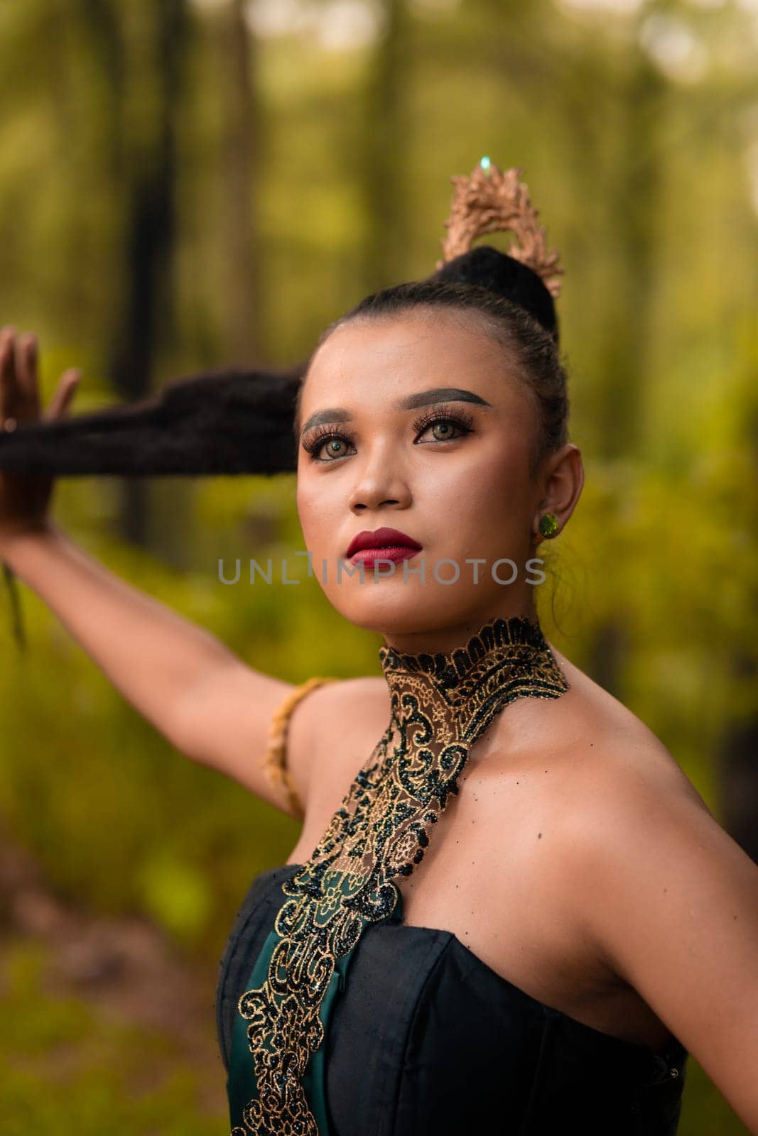 Gorgeous Asian woman in makeup with black hair and red lipstick wearing golden jewelry during the dance performance in front of the jungle