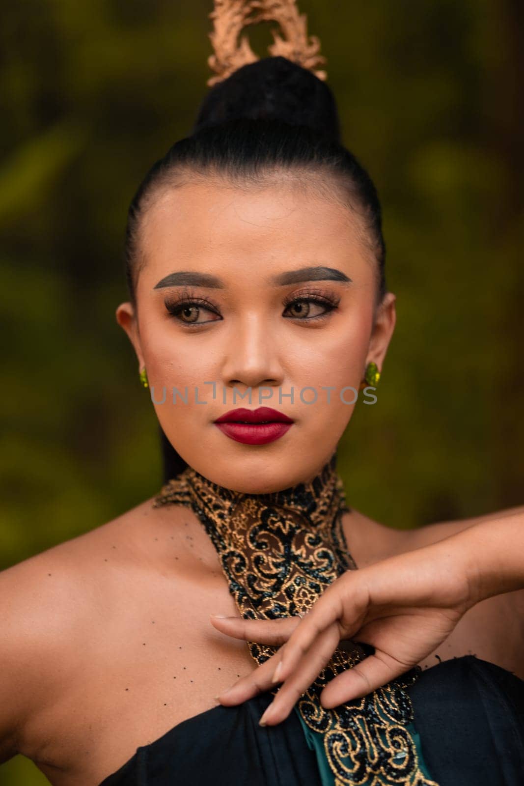 Portrait of an Asian woman in makeup and red lips while wearing a golden necklace and golden accessories on her black hair while standing in front of the jungle