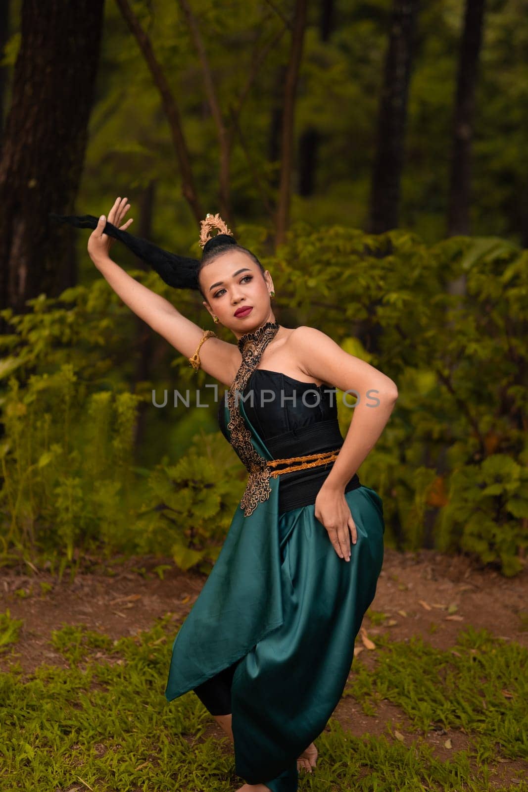 Asian woman holding her black hair in a green costume while wearing makeup and posing in front of the forest during the dance festival inside the village