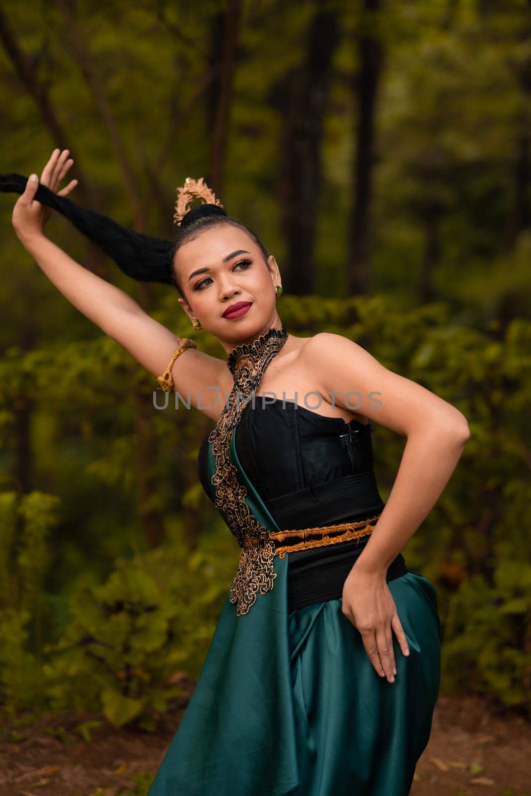 Asian woman holding her black hair in a green costume while wearing makeup and posing in front of the forest during the dance festival inside the village