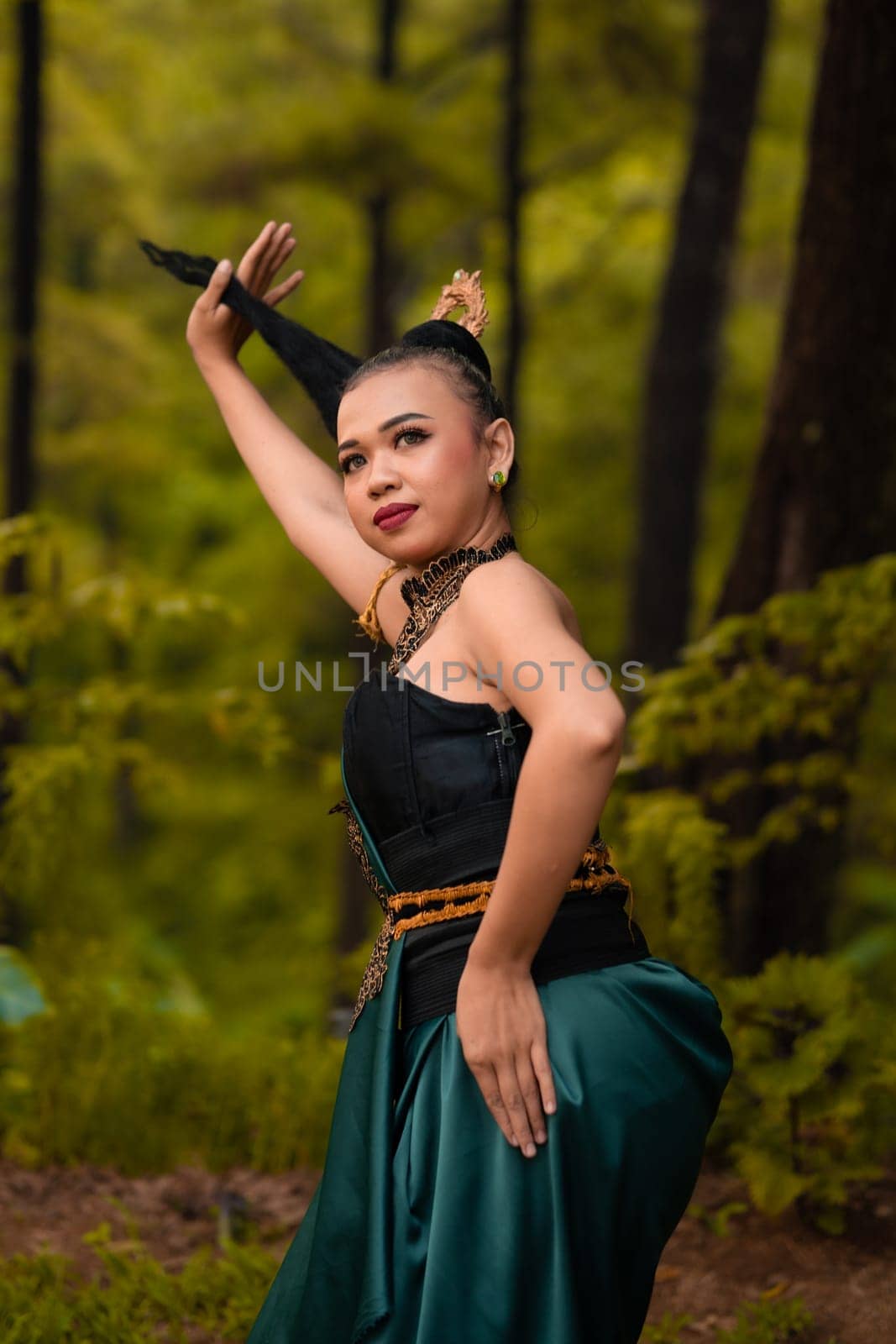 Asian woman holding her black hair in a green costume while wearing makeup and posing in front of the forest during the dance festival inside the village