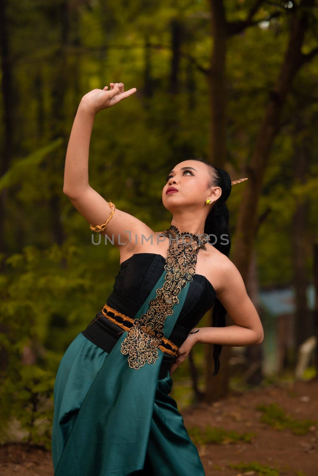 Balinese women raise their hands in the air while dancing in green costumes while performing in the festival in front of the jungle