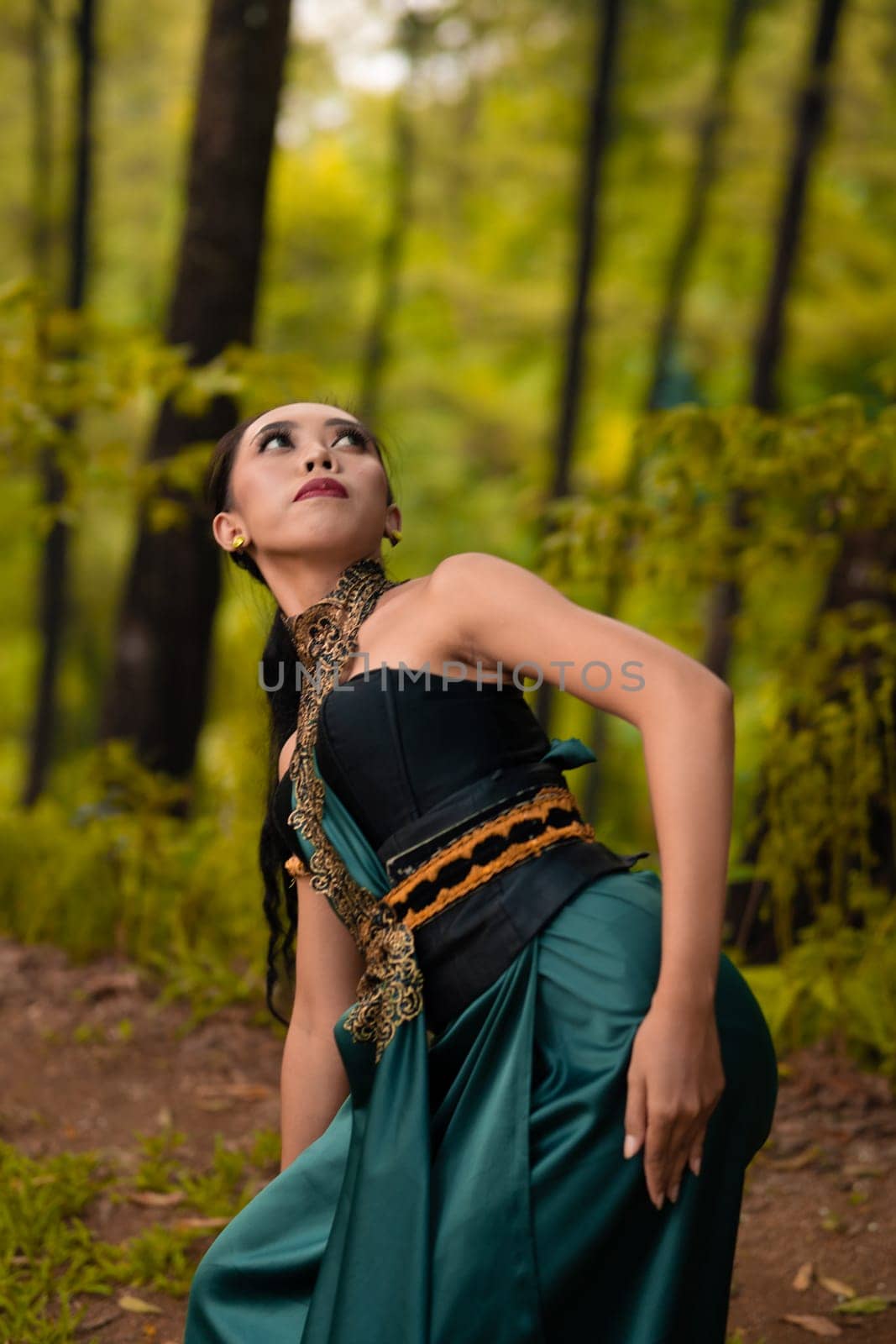 Beautiful Asian woman in a green costume with long black hair dances in the woods during the daylight
