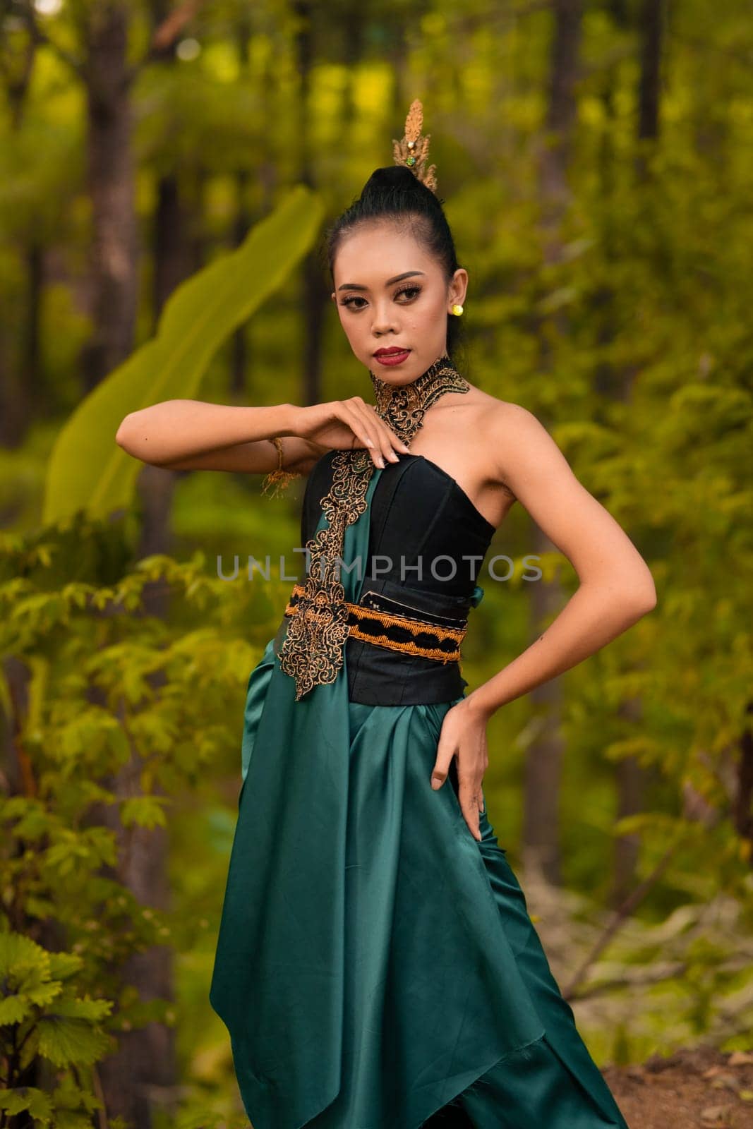 A brave Asian woman in makeup wearing a green costume and gold jewelry with the woods in the background during the morning
