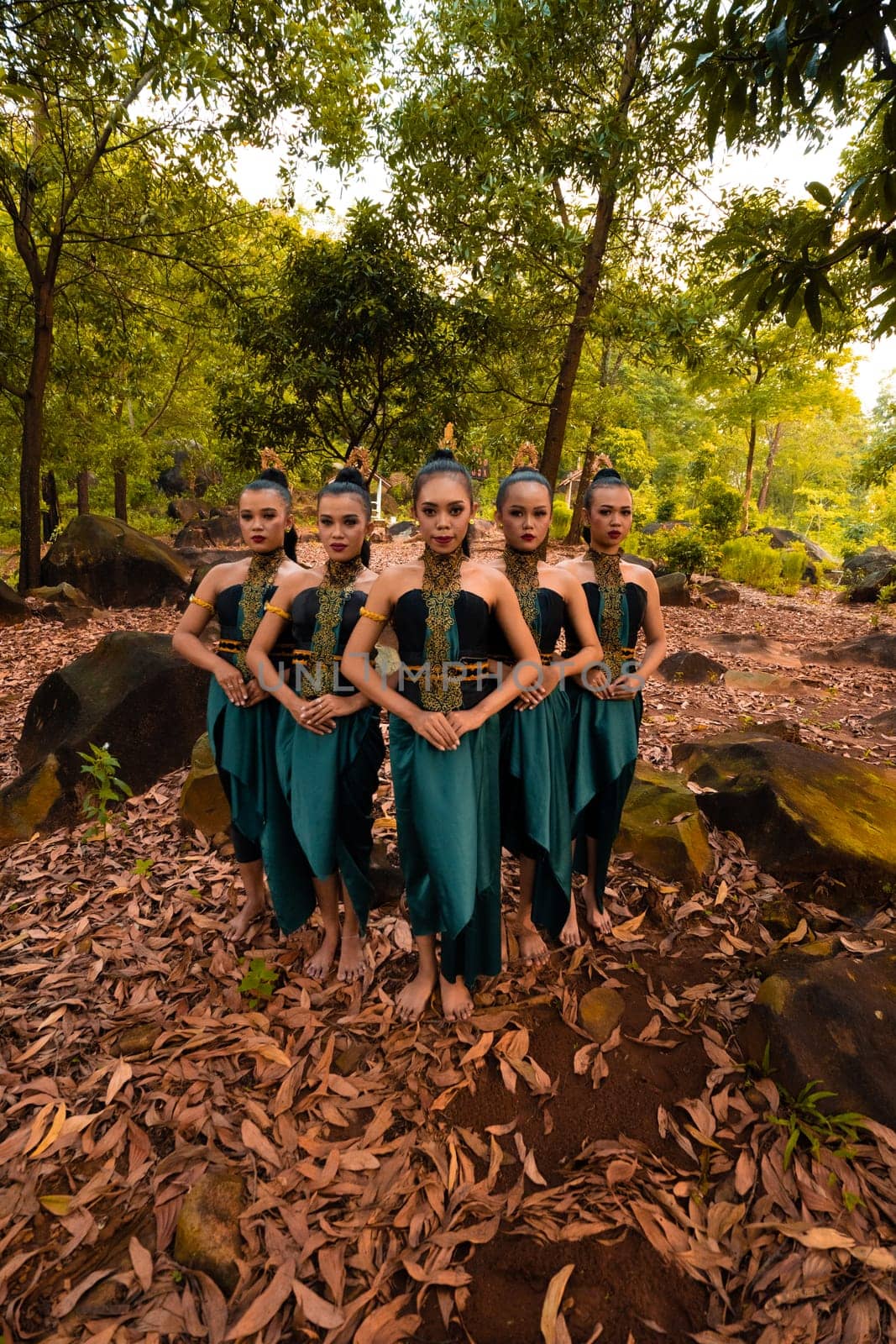 a wide shot of an Asian woman standing together in the forest while wearing a green dance costume with dead leaves in the background