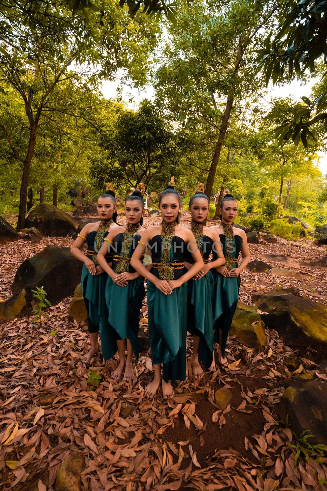 a wide shot of an Asian woman standing together in the forest while wearing a green dance costume with dead leaves in the background