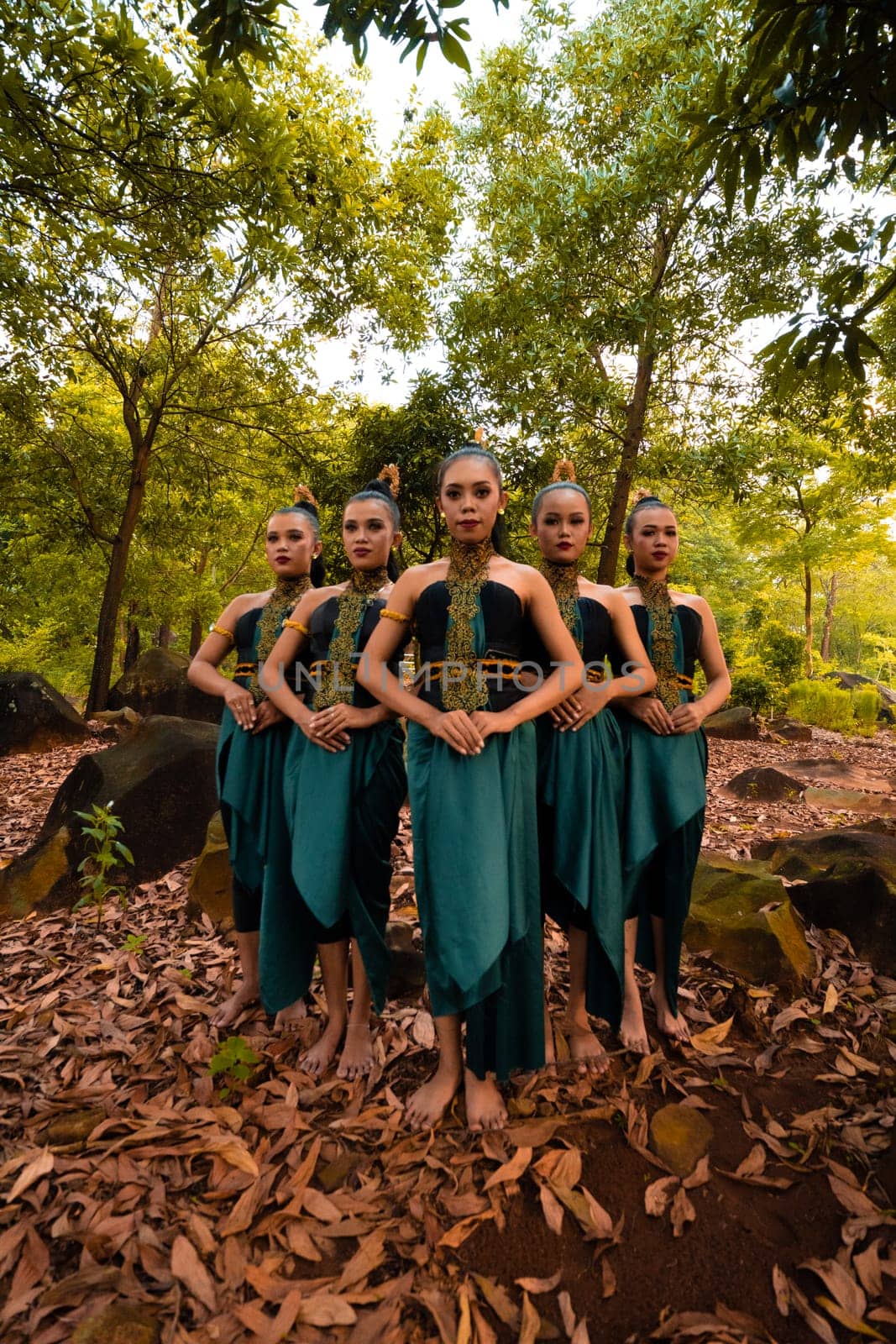 a wide shot of an Asian woman standing together in the forest while wearing a green dance costume with dead leaves in the background