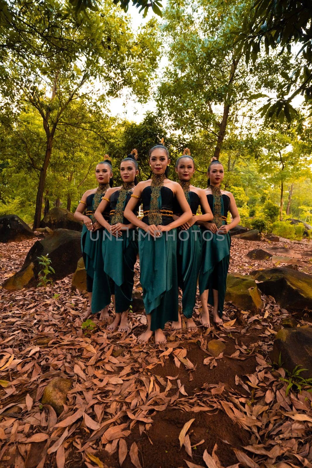 a wide shot of an Asian woman standing together in the forest while wearing a green dance costume with dead leaves in the background