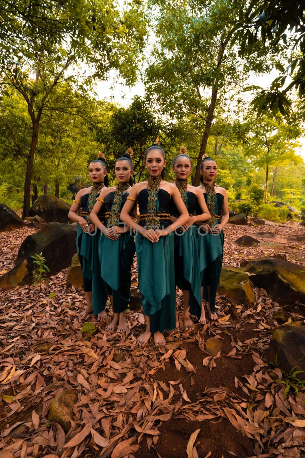 a wide shot of an Asian woman standing together in the forest while wearing a green dance costume with dead leaves in the background