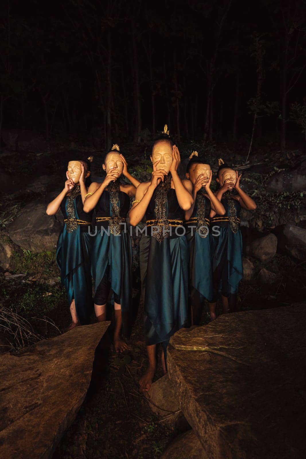 A Group of Asian women wearing a wooden masks in green costumes while standing between the rocks inside the forest during the night