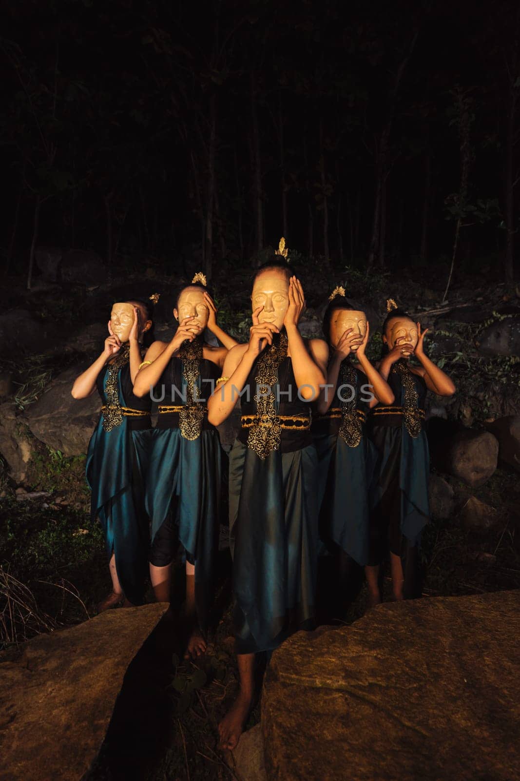 A Group of Asian women wearing a wooden masks in green costumes while standing between the rocks inside the forest during the night