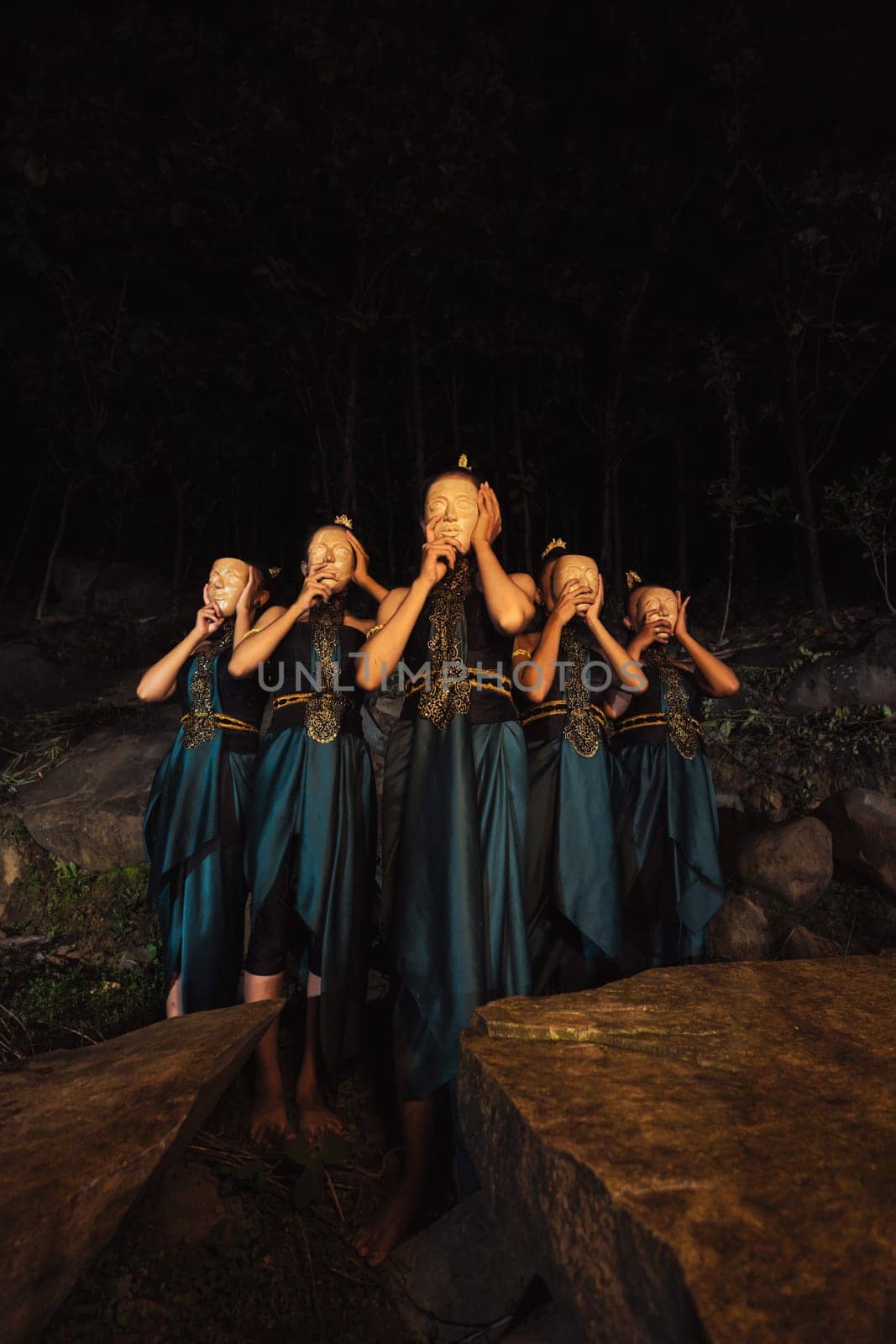 A Group of Indonesian women holding and wearing a brown wooden mask in a green skirt between the rock inside the woods