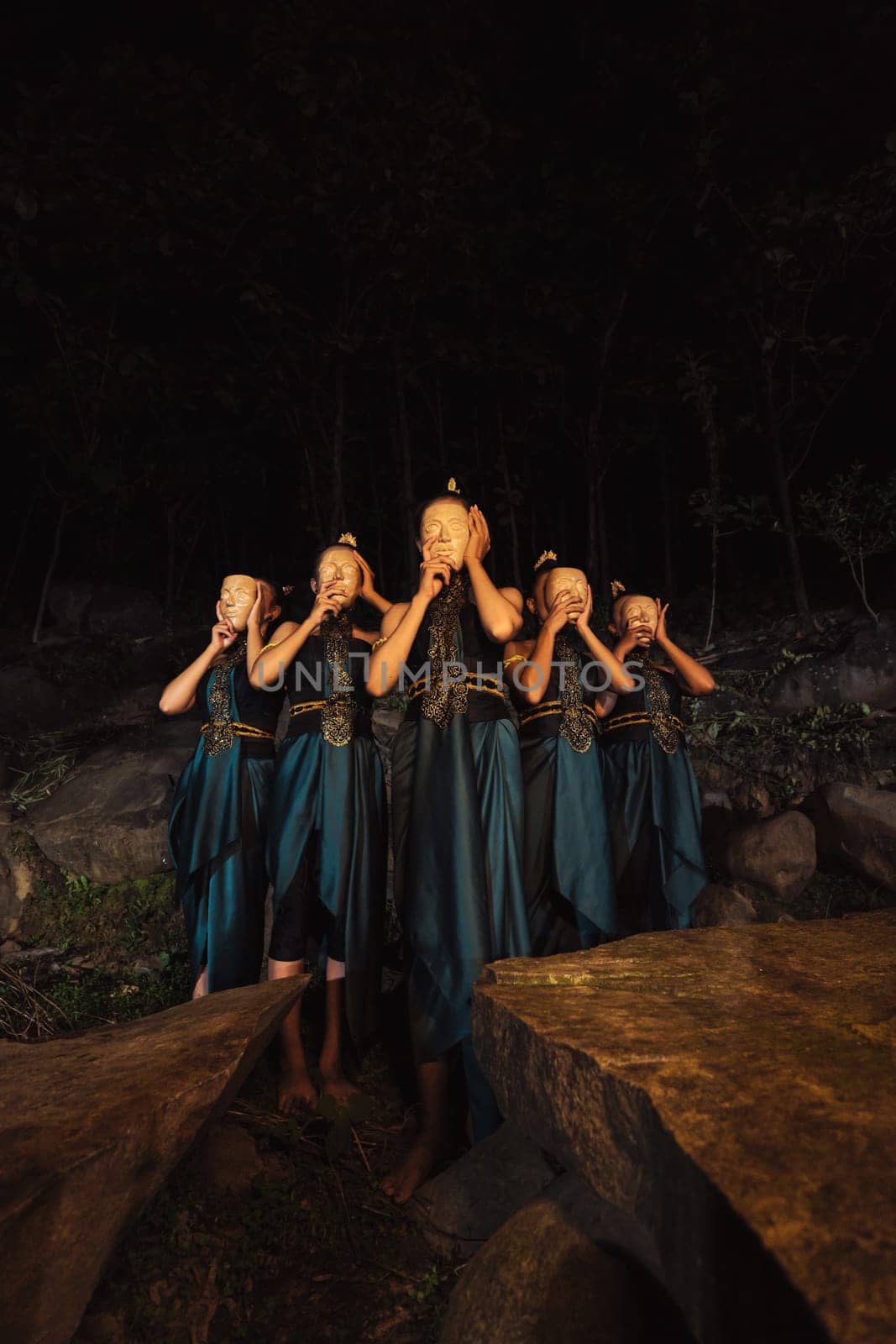 A Group of Indonesian women holding and wearing a brown wooden mask in a green skirt between the rock inside the woods