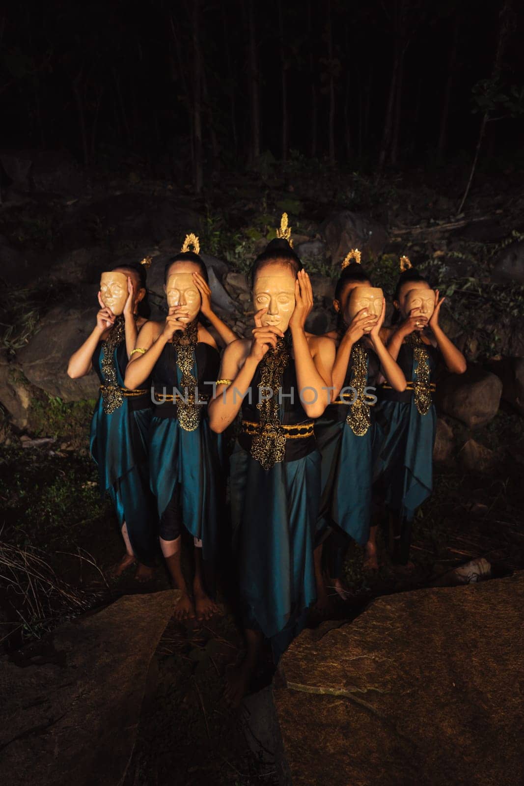 A Group of Asian women wearing a wooden masks in green costumes while standing between the rocks inside the forest during the night