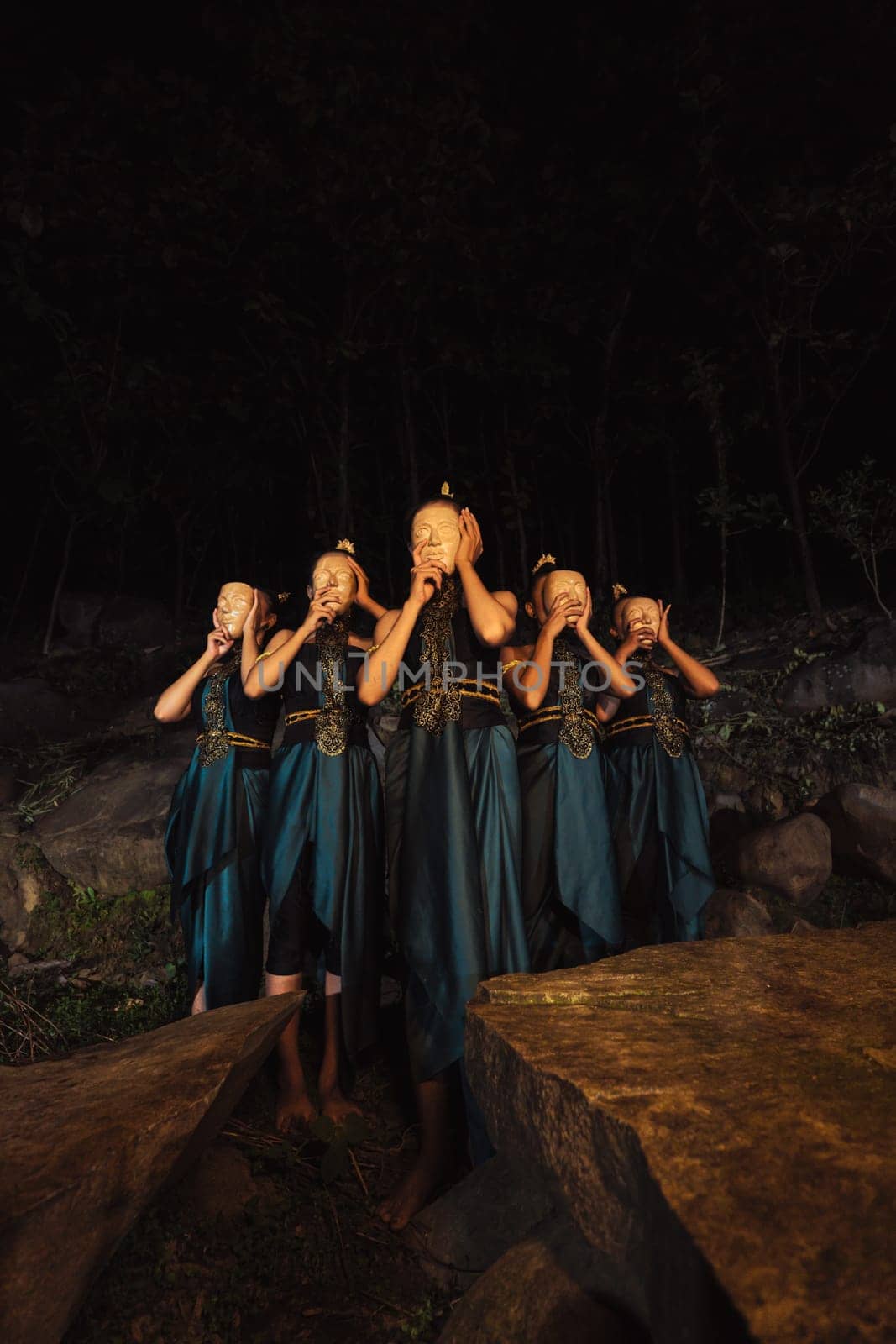 A Group of Indonesian women holding and wearing a brown wooden mask in a green skirt between the rock inside the woods