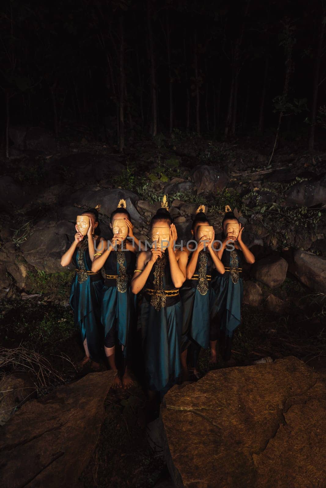 A Group of Indonesian women holding and wearing a brown wooden mask in a green skirt between the rock inside the woods