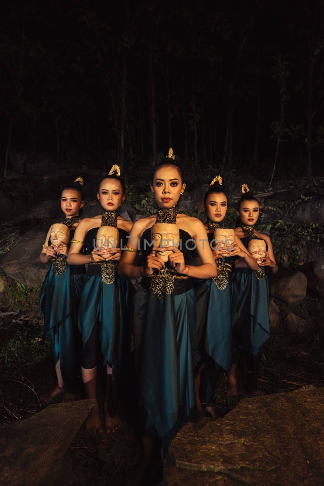 A Bunch of girls in green costumes holding a wooden masks in their hands while standing between the big rocks in front of the forest