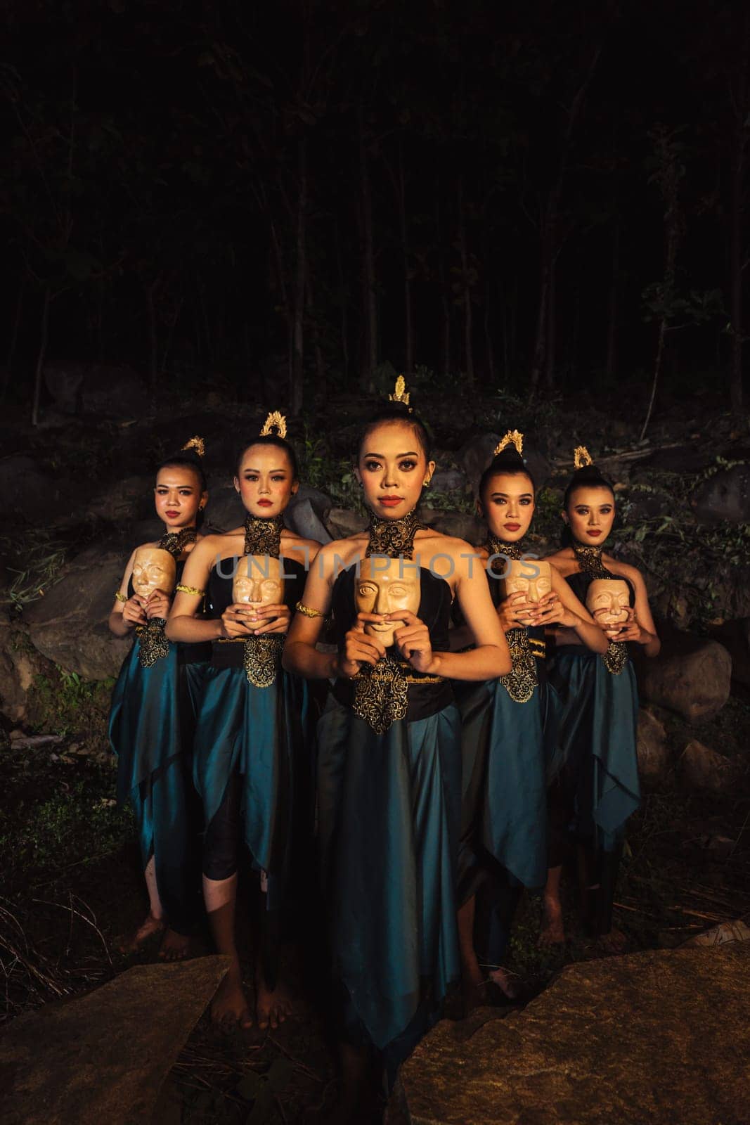 A Bunch of girls in green costumes holding a wooden masks in their hands while standing between the big rocks in front of the forest