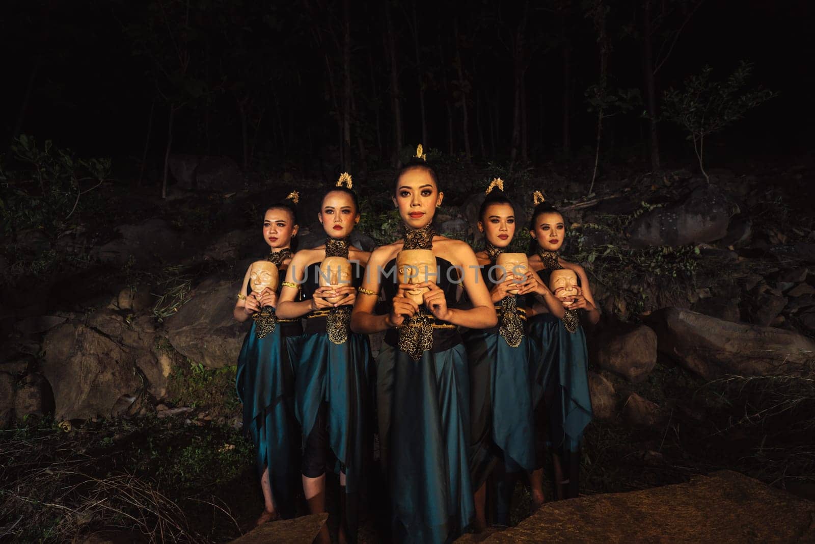 A Group of ethnical women holding a wooden craft mask in their hands while in line inside the wood in the night