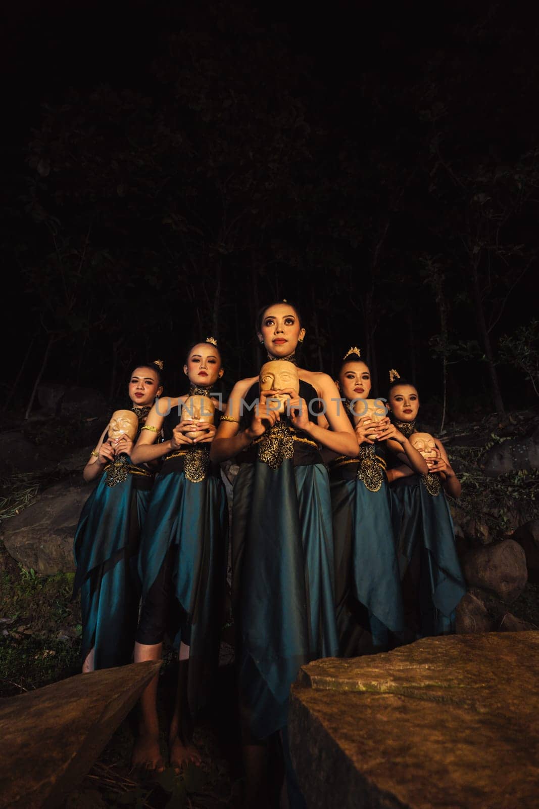 A Bunch of girls in green costumes holding a wooden masks in their hands while standing between the big rocks in front of the forest