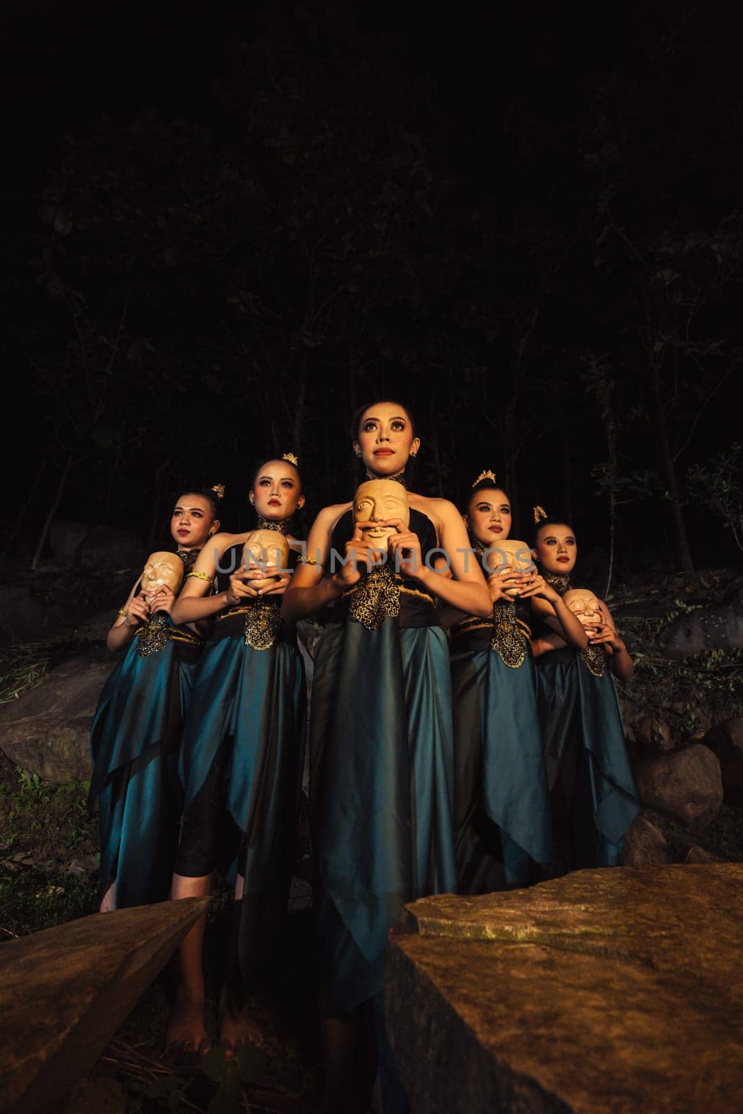 A Bunch of girls in green costumes holding a wooden masks in their hands while standing between the big rocks in front of the forest