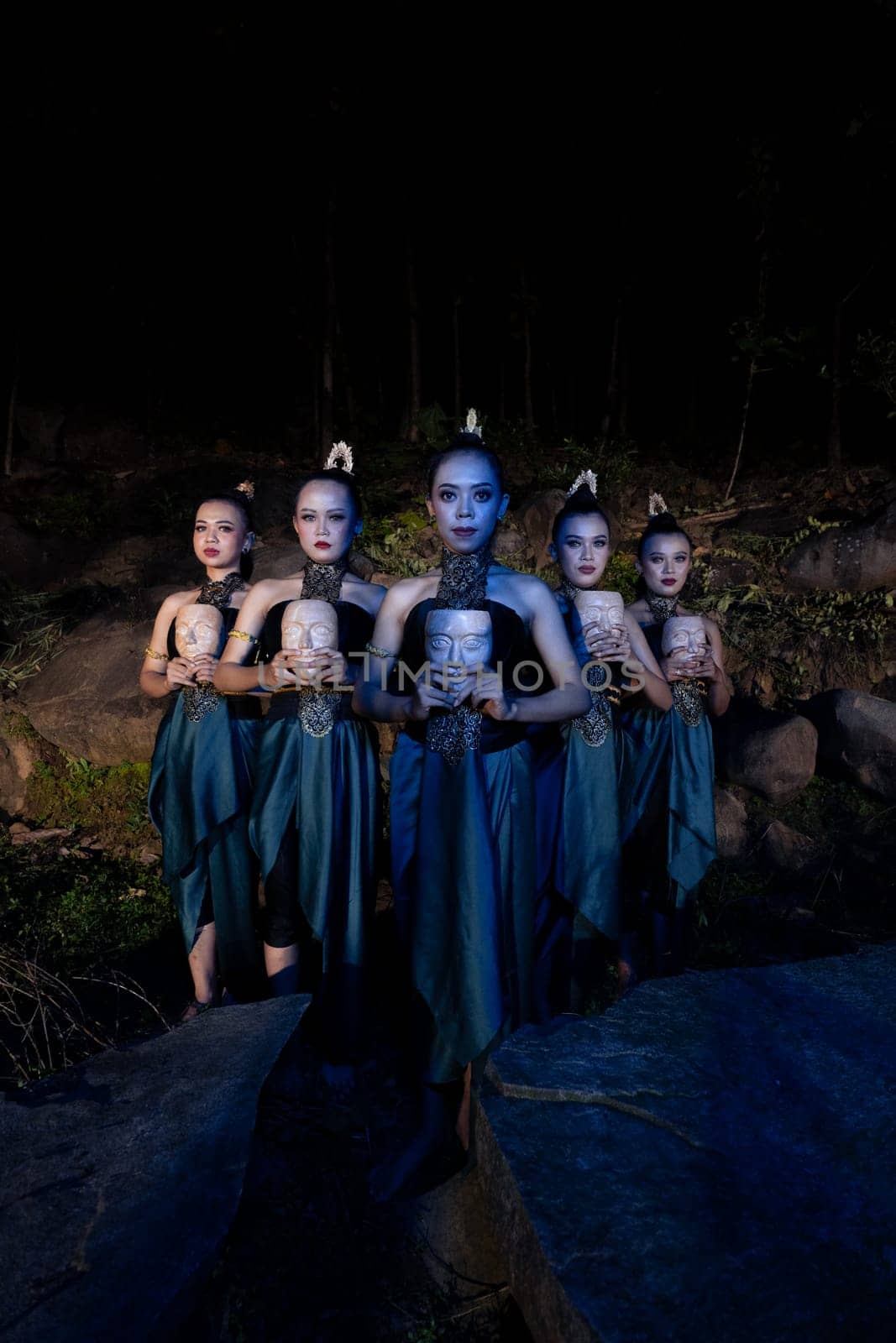 A Group of Javanese women standing in the woods between the rocks while holding a wooden mask inside the village