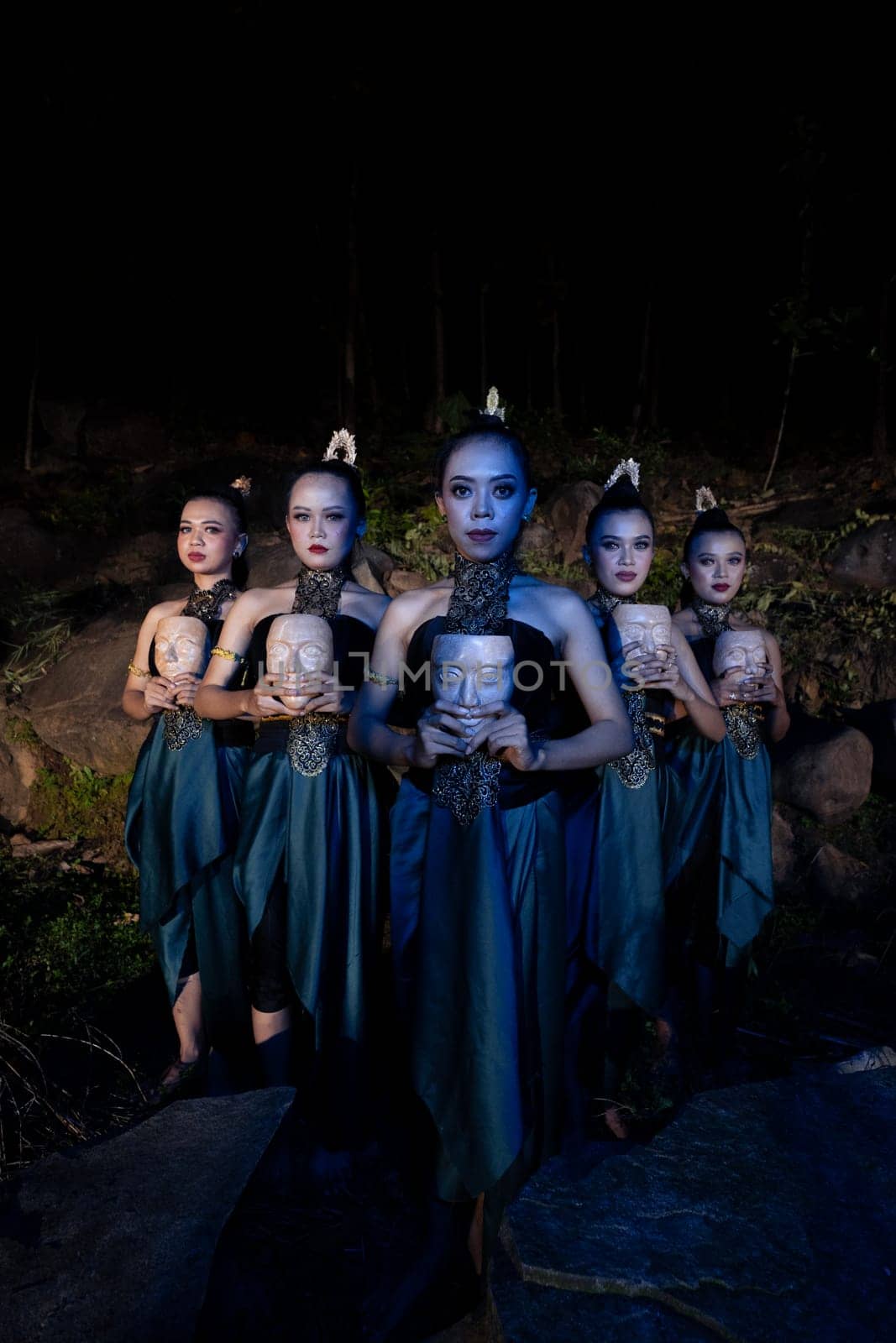 A Group of Javanese women standing in the woods between the rocks while holding a wooden mask inside the village