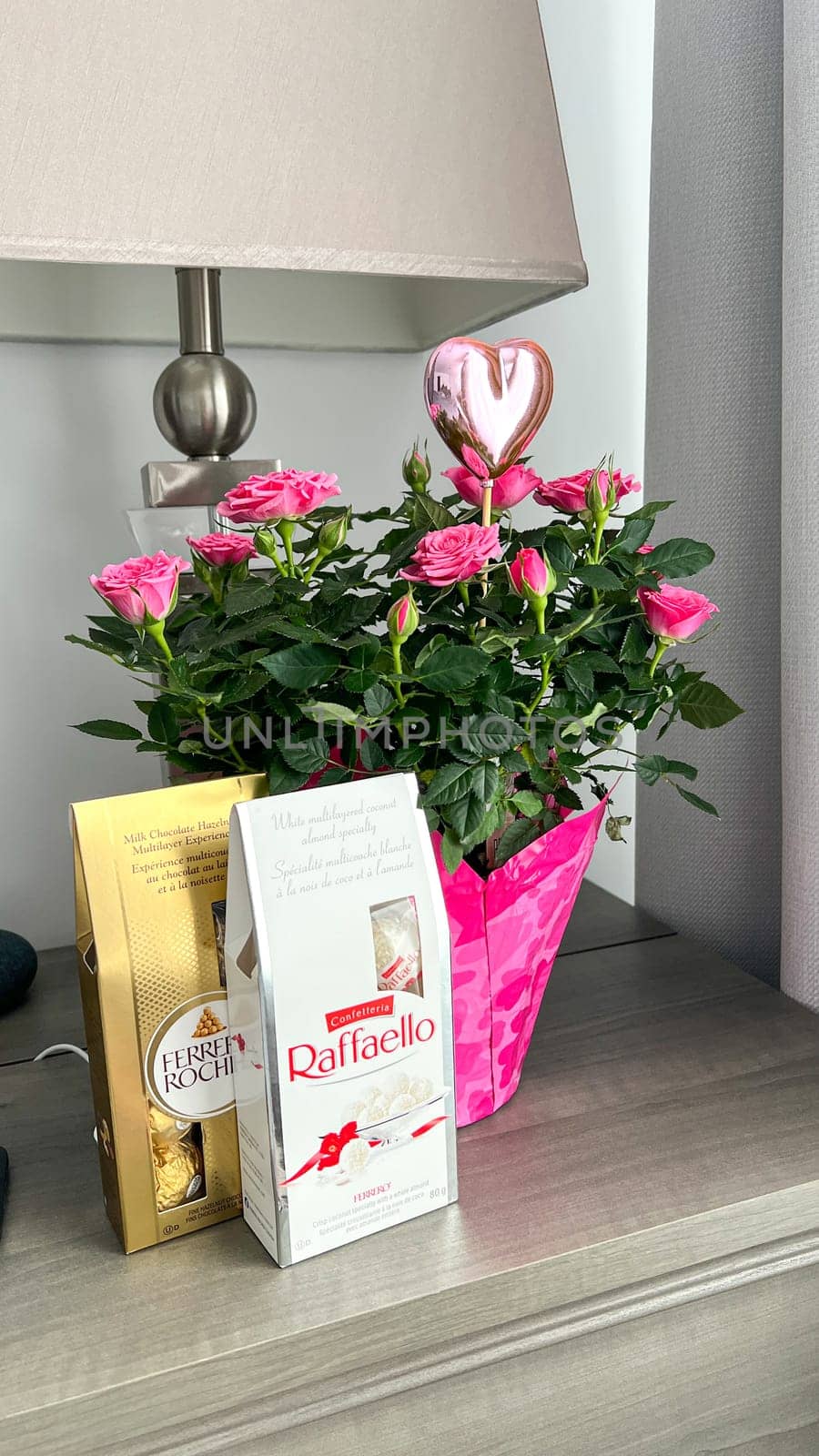 Ottawa, Canada - March 01, 2025: Pink roses in a decorative pot with a heart-shaped balloon and Raffaello, Ferrero Rocher chocolates on a wooden table, creating a romantic atmosphere.