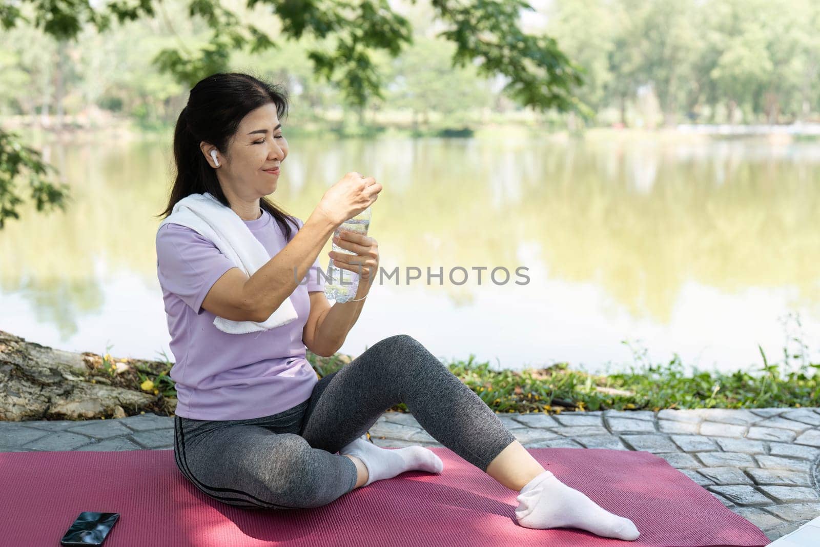 A focused woman sits on a yoga mat, ready to drink water after her outdoor yoga session by the tranquil lake.