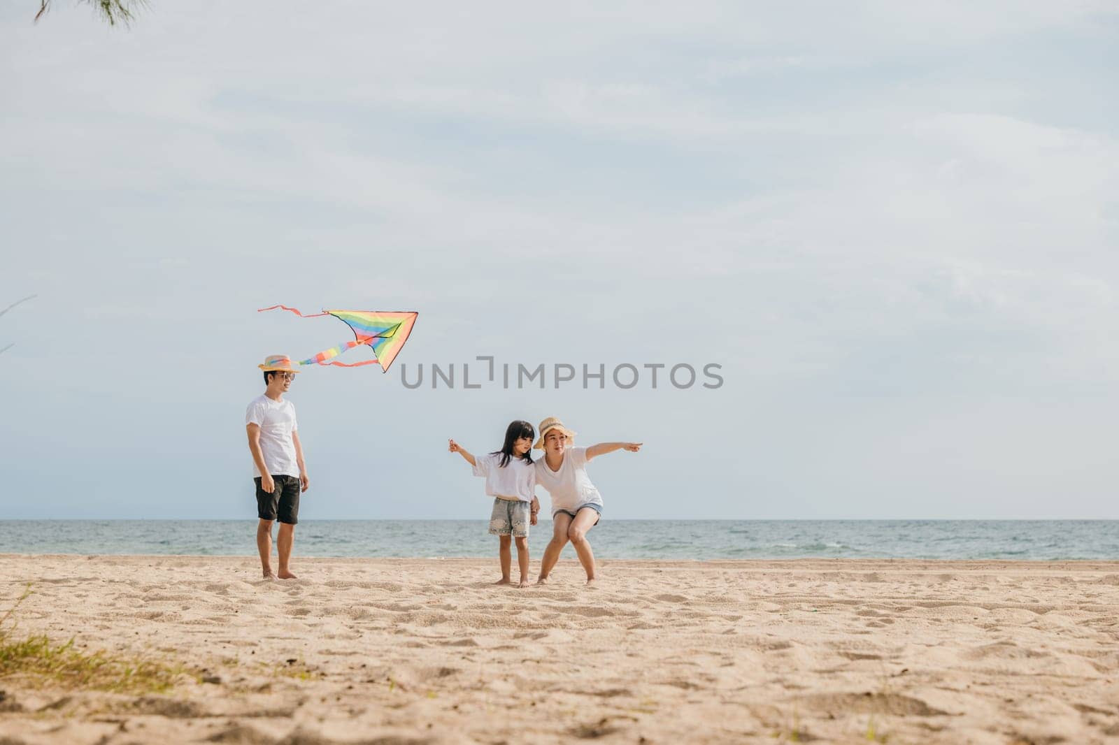 Asian family is flying a kite high in the sky while running on the beach. Happy parents mother and father with their child playing with kite, family is enjoying their time together, summer day