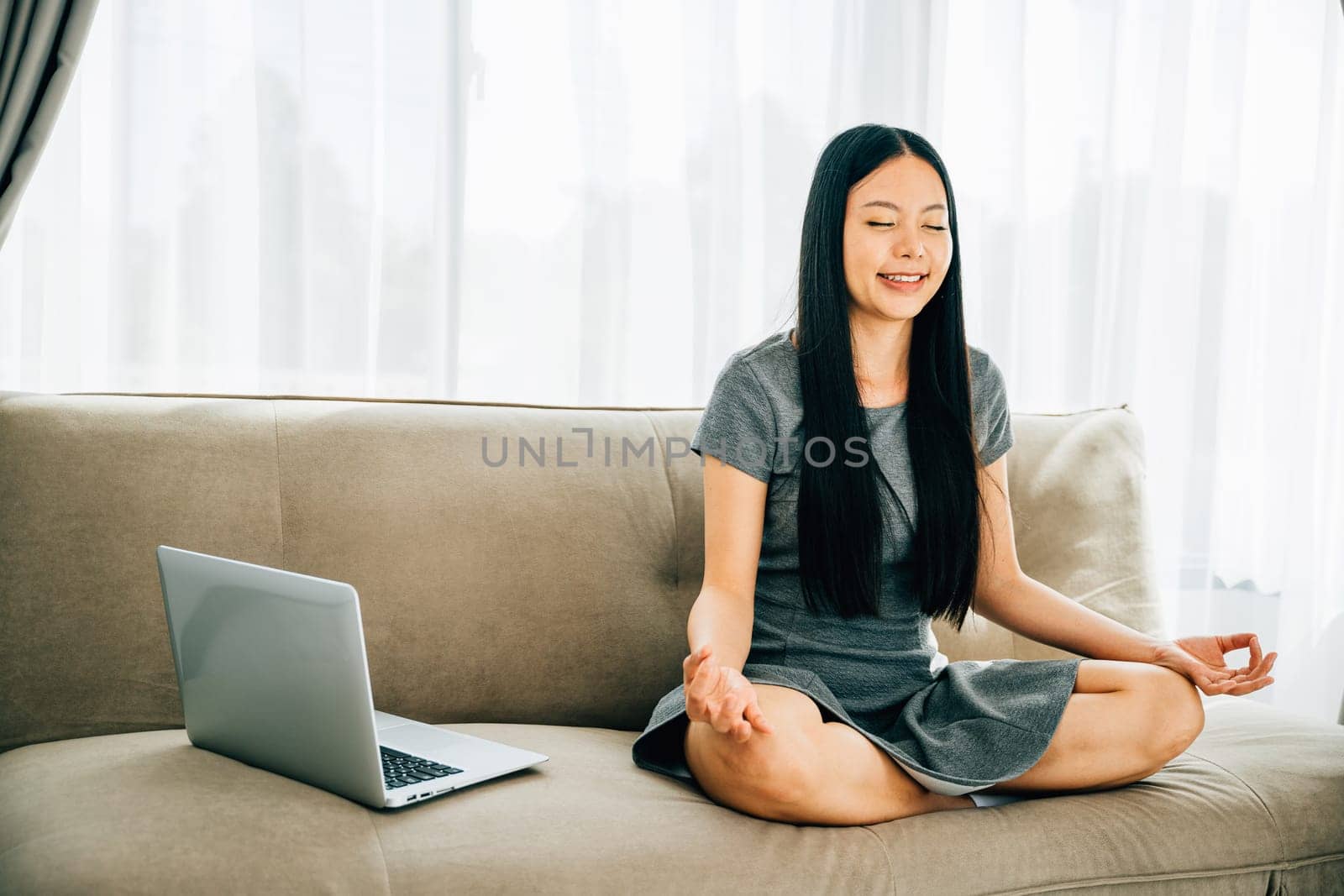 Asian woman finds balance meditating in lotus pose with laptop on sofa. Serene businesswoman practices relaxation and mindfulness while working online. Portrait of a smiling student in harmony.