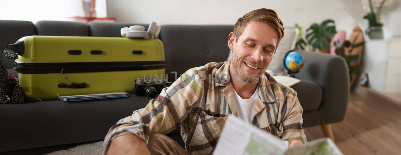 Portrait of handsome smiling man, holding map, sitting with suitcase in living room, going abroad on holiday, studying his route. Tourism concept