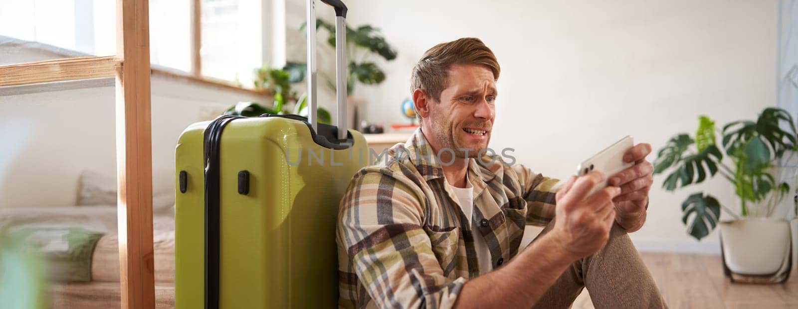 Portrait of stressed man looks at his mobile phone, playing game on smartphone, sitting near suitcase, waiting for his flight or taxi. Travelling concept
